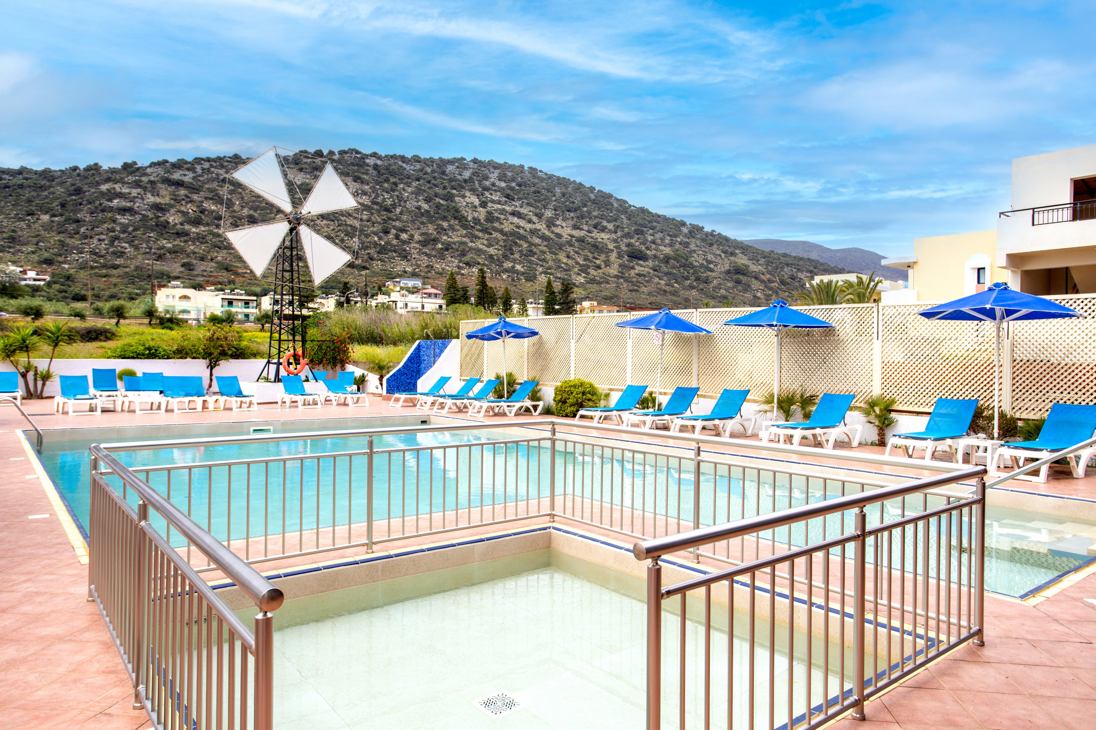 An outdoor resort swimming pool is surrounded by blue sun loungers and matching umbrellas. A shallow fenced pool area sits in the foreground, suggesting a children’s or wading section. In the background, a white windmill structure and safety ring add visual interest near the pool deck. Beyond the resort wall, green fields and a tree-covered hill rise under a bright blue sky with light clouds.