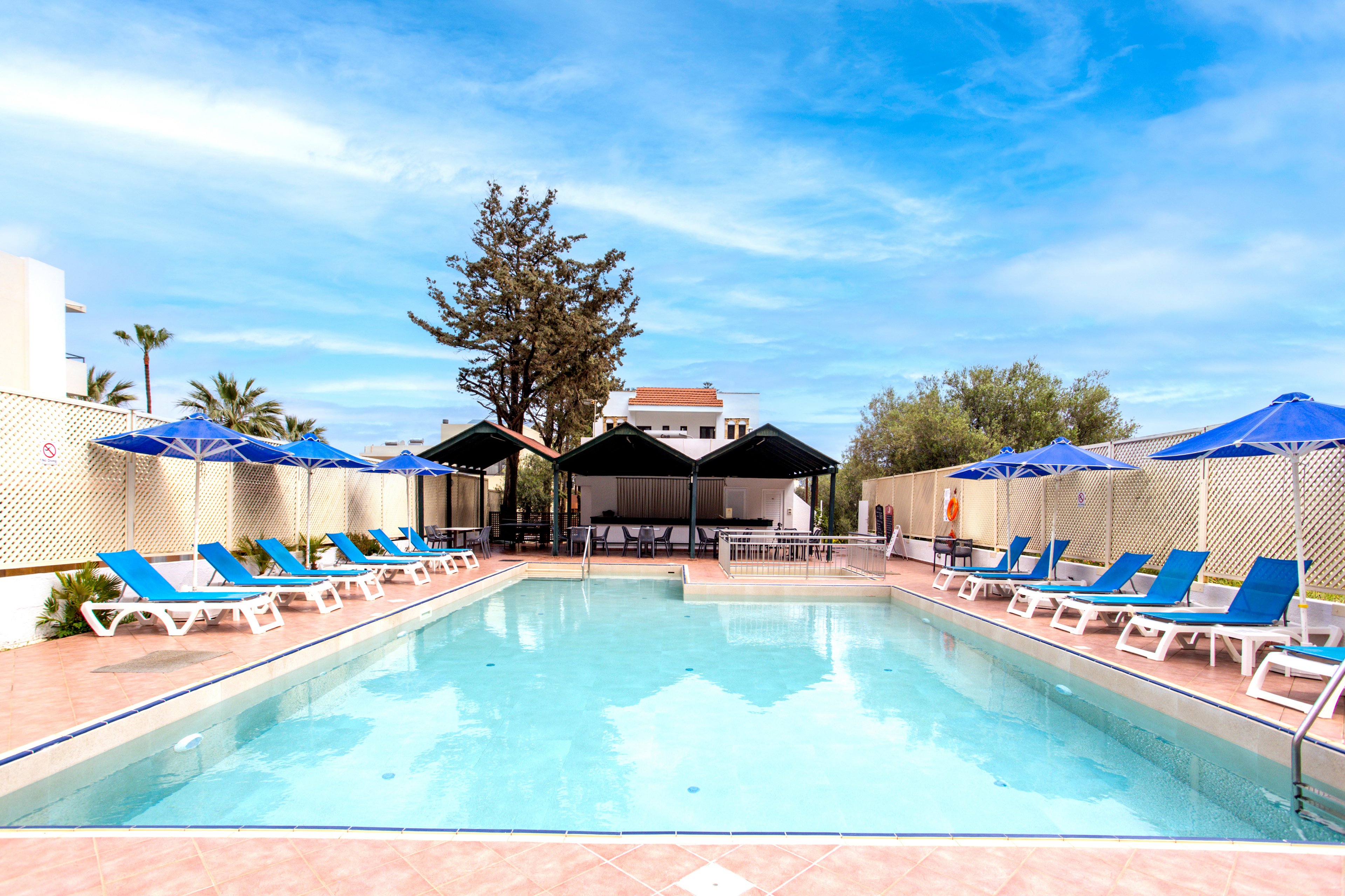 A clear blue swimming pool is surrounded by rows of sun loungers and umbrellas in a resort setting. The pool area is framed by light-colored tiles and lattice fencing, creating a clean and organized look. Blue umbrellas and cushions add a vibrant contrast against the pale deck and bright sky. In the background, small buildings and trees suggest a warm vacation destination.