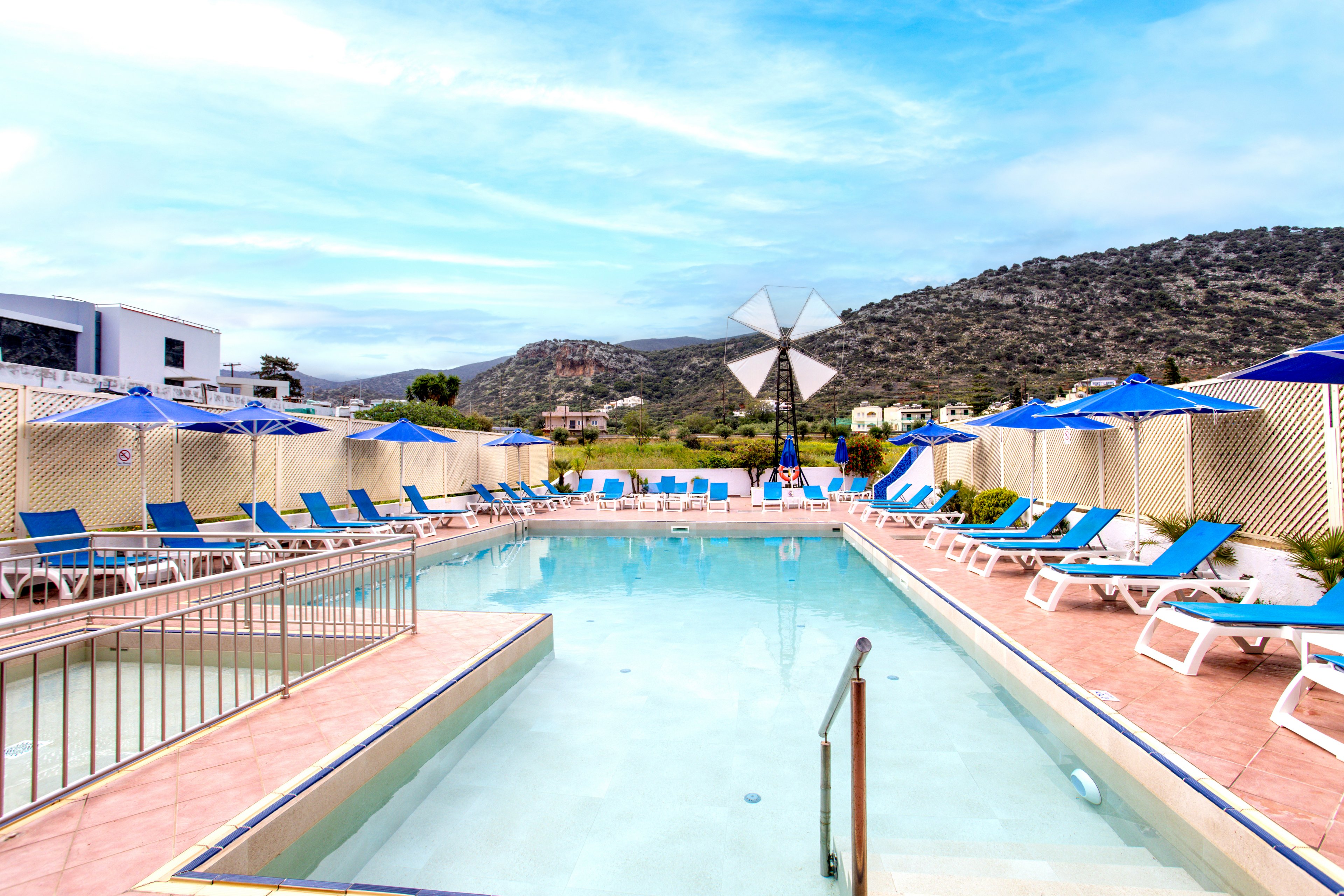 A bright outdoor swimming pool is surrounded by blue sun loungers and umbrellas at a resort. The pool area is enclosed by light-colored fencing, creating a private and relaxing atmosphere. In the background, a white windmill structure stands against rolling hills and scattered buildings. The sky is clear with soft clouds, enhancing the tranquil vacation setting.