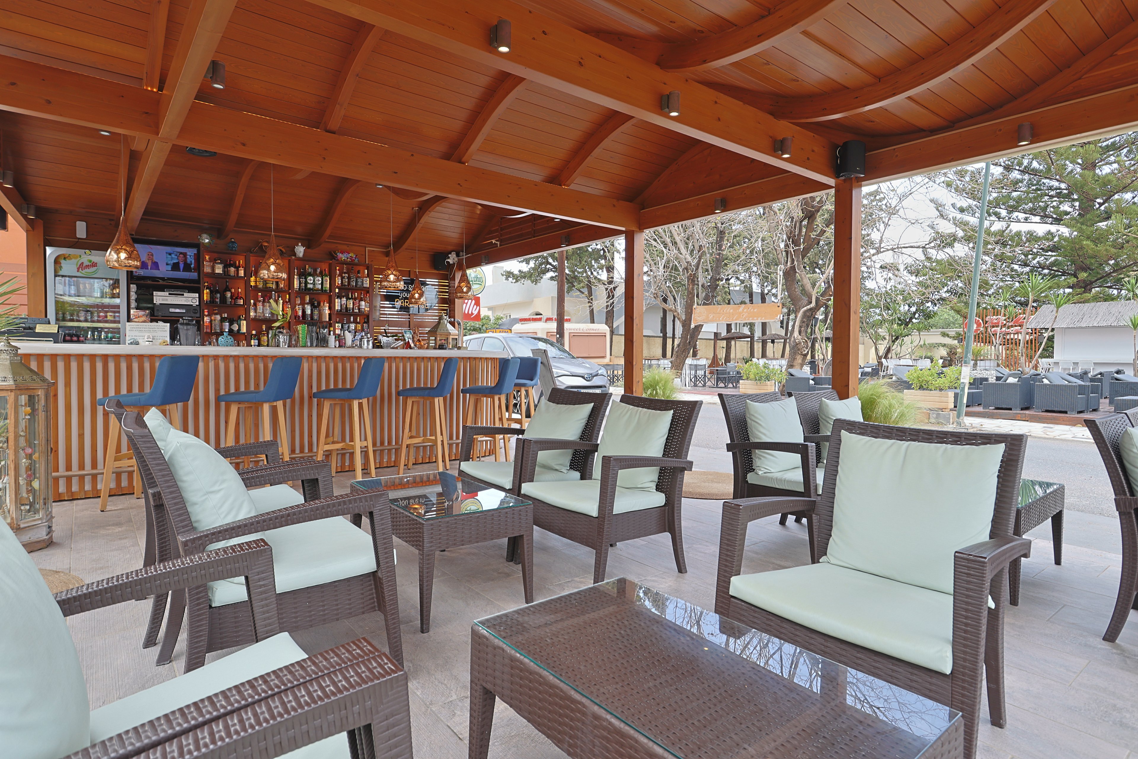 An open-air bar with a wooden roof features a row of blue barstools facing a fully stocked counter. In the foreground, wicker lounge chairs with light cushions surround glass-topped tables, creating a relaxed seating area. The setting appears to be a poolside or resort patio with trees and additional outdoor furniture in the background. The overall scene suggests a casual, social atmosphere for guests to enjoy drinks and conversation.