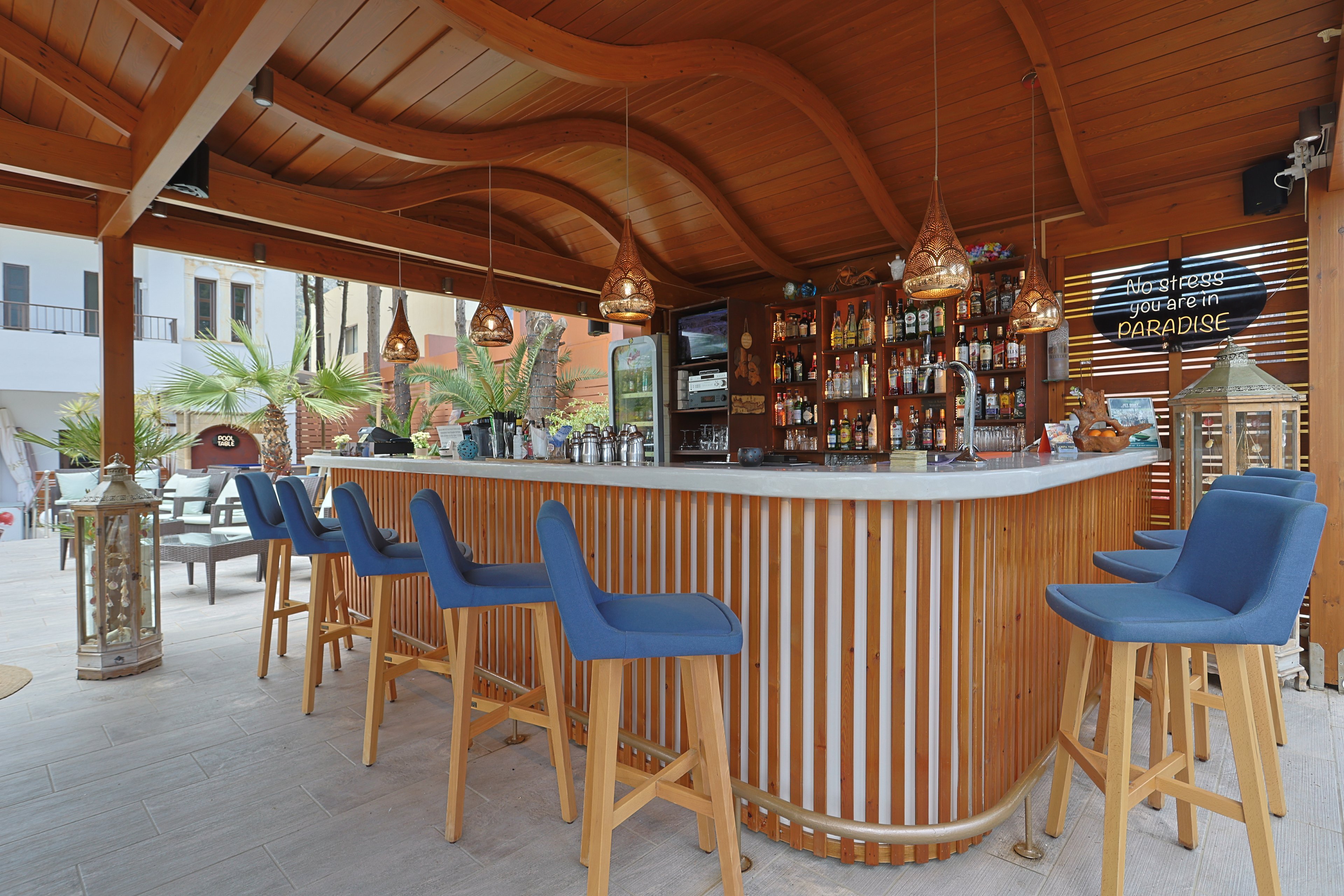 An open-air bar with a curved wooden counter and blue cushioned bar stools stands under a sculpted wooden ceiling. The back bar is stocked with various bottles and glassware, framed by warm pendant lights. Palm trees and resort buildings are visible in the background, suggesting a tropical or coastal setting. The overall scene feels relaxed and inviting, ideal for vacation or leisure time.