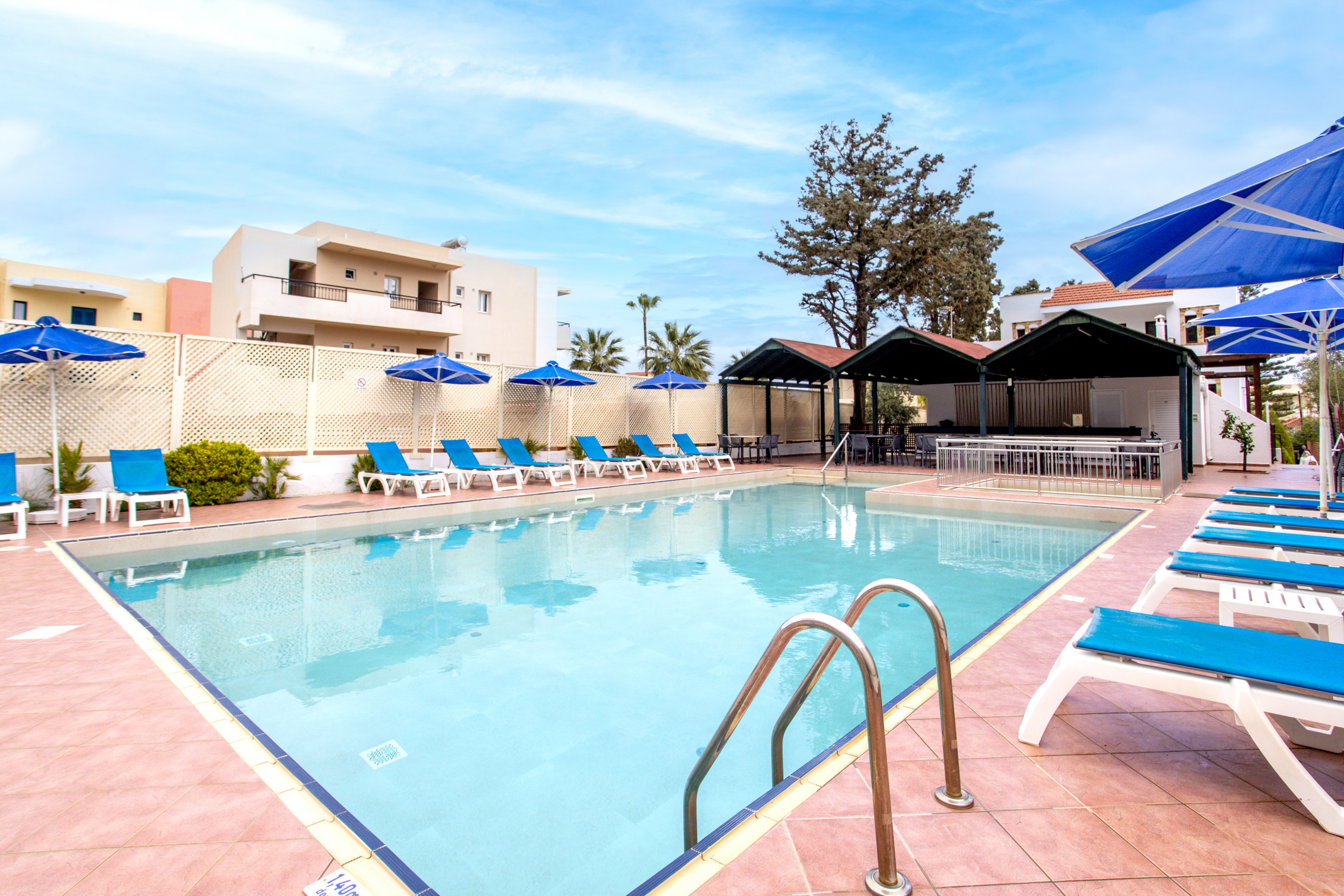 An outdoor hotel swimming pool is surrounded by blue sun loungers and umbrellas on a tiled deck. Metal handrails lead into the clear water, reflecting the bright sky above. In the background, low-rise modern buildings and a poolside bar area create a relaxed resort atmosphere. The scene suggests a warm, sunny day ideal for vacation and leisure.