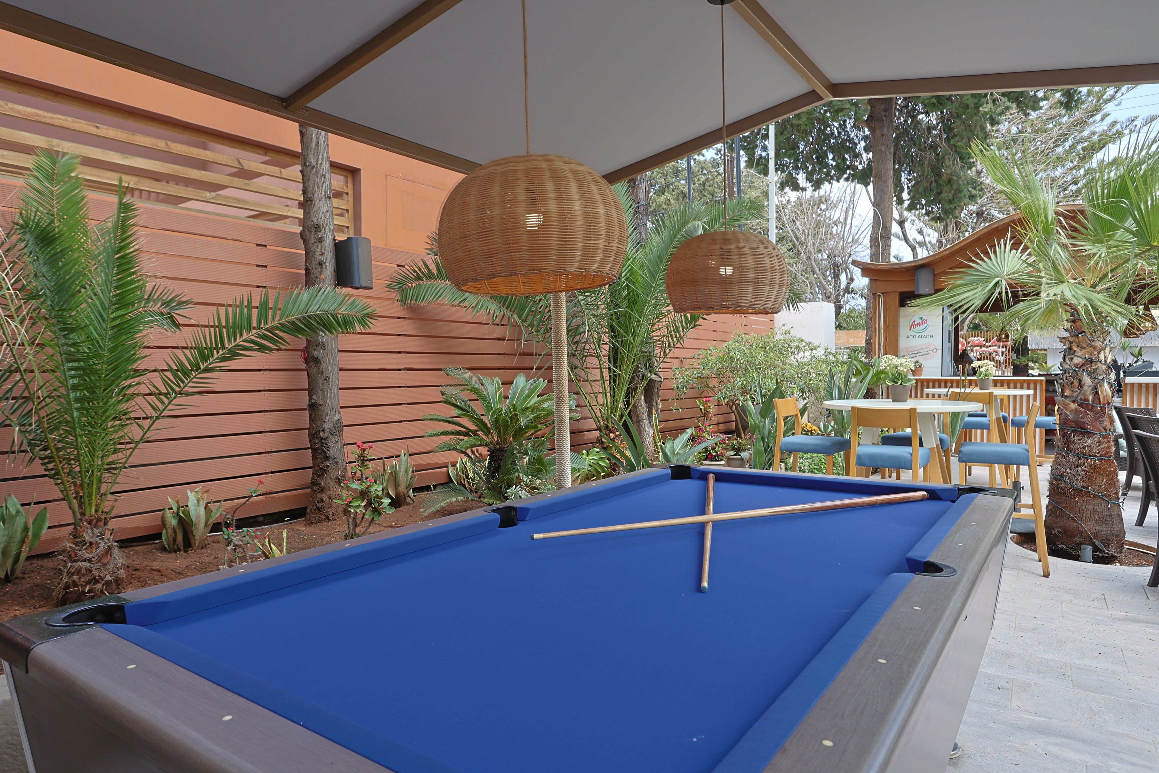 An outdoor recreation area features a blue felt pool table under a shaded canopy. Two pool cues rest crossed on the table, ready for a game. In the background, there is a seating area with tables and chairs, surrounded by palm plants and wooden fencing. Warm hanging lamps and greenery create a relaxed, casual atmosphere.