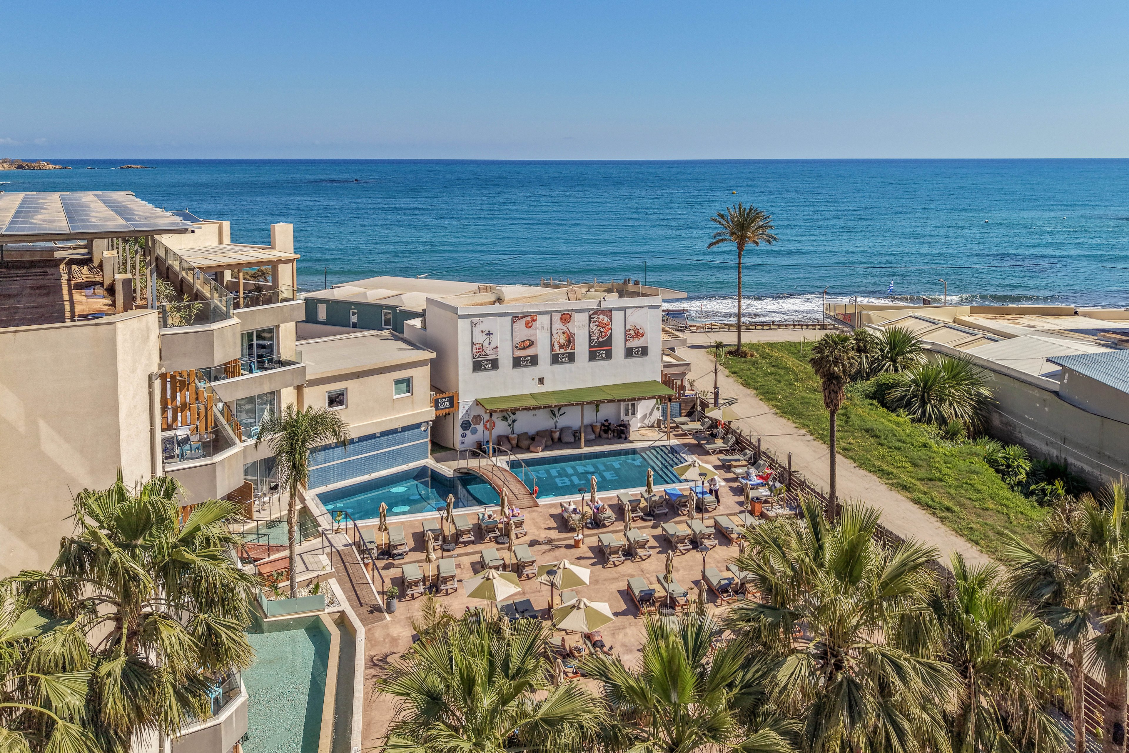 A beachfront resort features a central swimming pool surrounded by numerous sun loungers and umbrellas. Guests relax on the pool deck while others enjoy the bar and shaded seating areas. The hotel buildings frame the pool, with balconies overlooking the scene and palm trees adding a tropical feel. In the background, calm blue ocean waves meet a sandy shoreline under a clear sky.