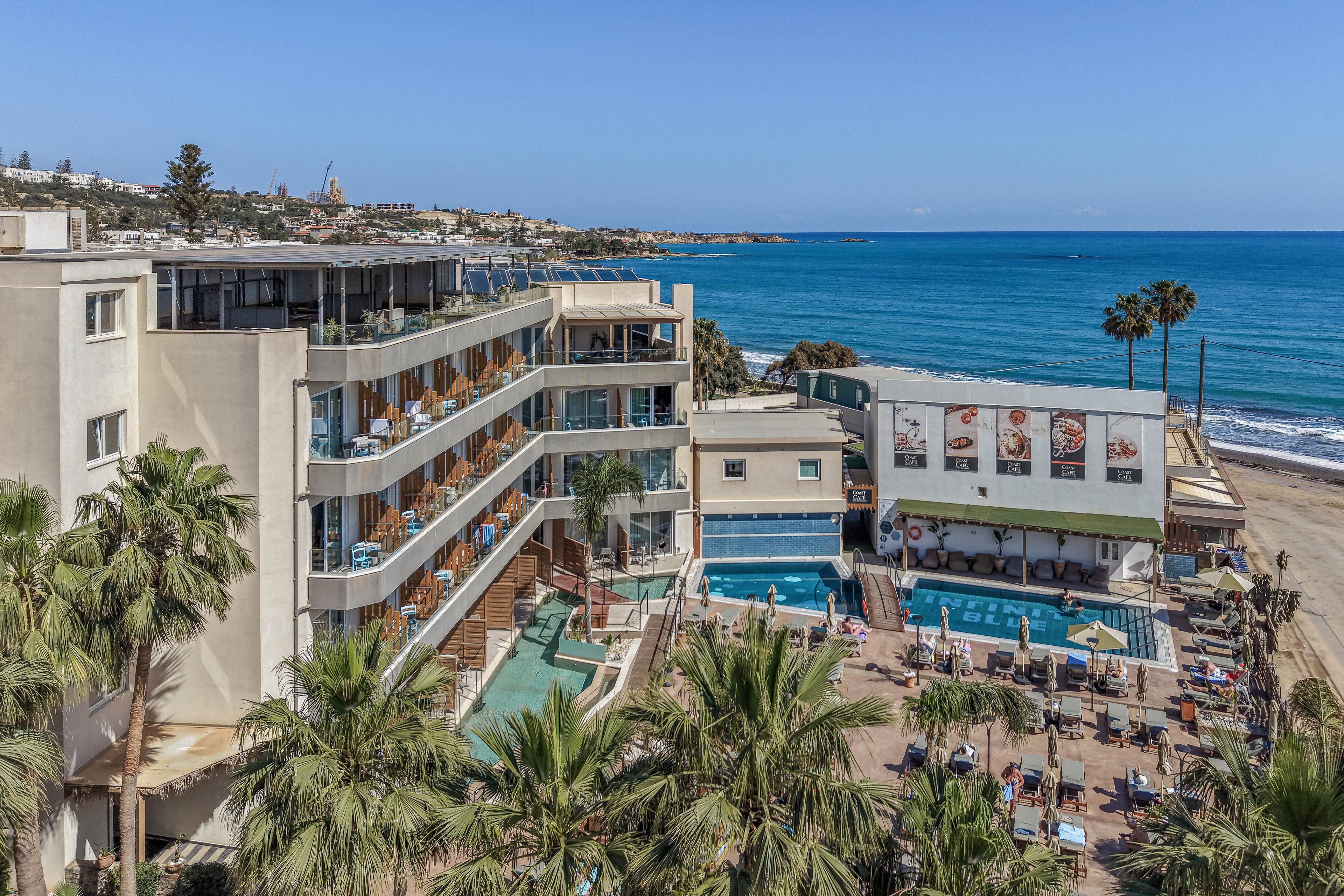 A multi-story beachfront hotel overlooks a bright blue ocean and sandy shore. Guests relax on balconies and around an outdoor swimming pool beside the sea. Palm trees and sun loungers create a classic coastal resort atmosphere under a clear sky. Large posters decorate the side of a nearby building, adding visual interest to the scene.