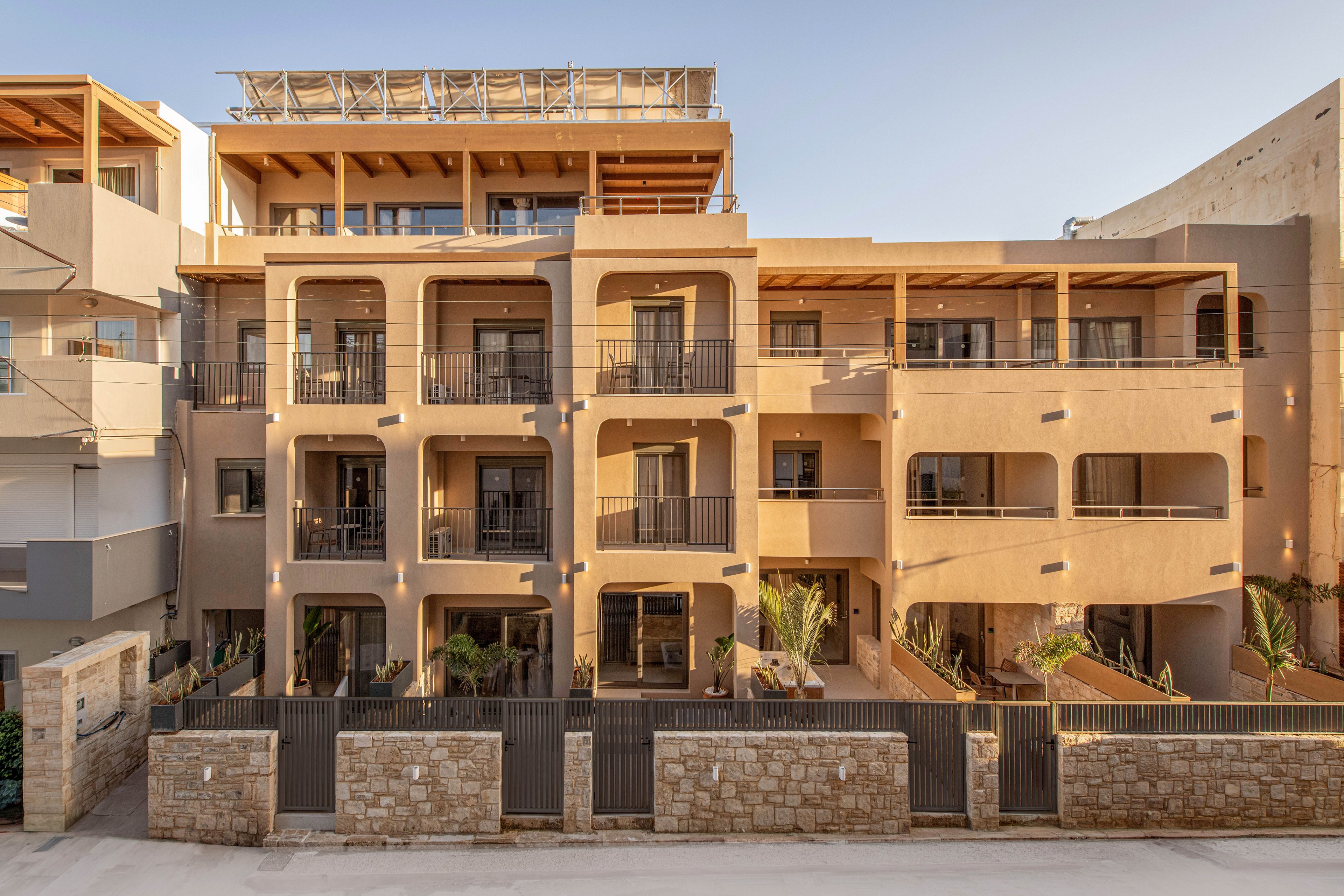 A contemporary multi-story apartment building is shown in warm evening light. The facade features repeating arches, balconies with dark metal railings, and a neutral beige color palette. A low stone wall and metal gates line the front of the property, separating it from the street. Adjacent structures are partially visible on both sides, emphasizing the urban residential setting.