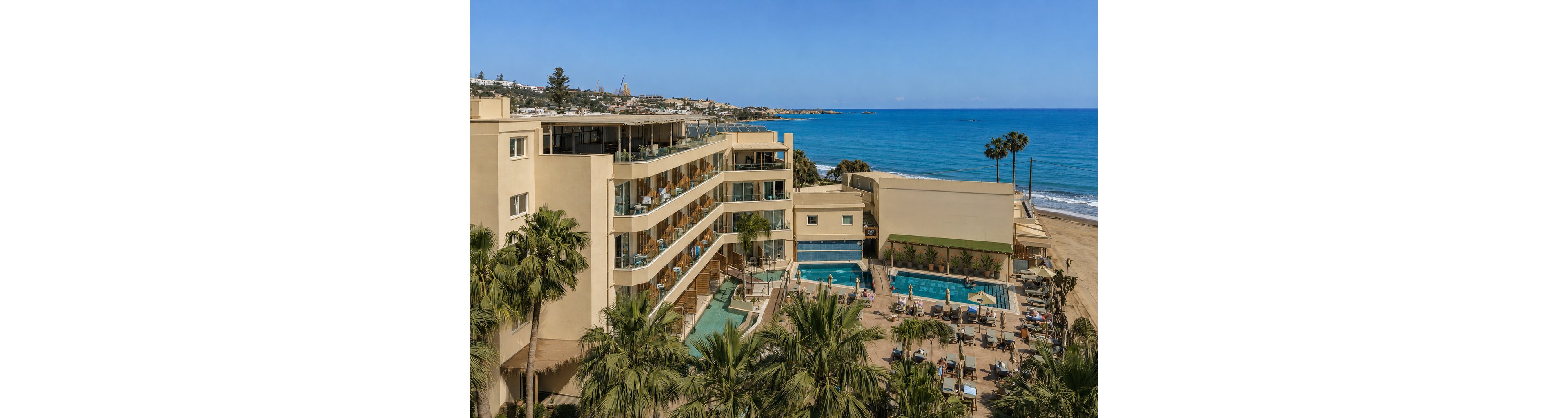 A multi-story beachfront resort overlooks a sandy shoreline and bright blue sea. Several outdoor swimming pools and sun loungers are arranged around the hotel, with guests relaxing in the sun. Palm trees frame the scene, adding a tropical feel to the coastal setting. The clear sky and calm water suggest a warm, sunny vacation destination.