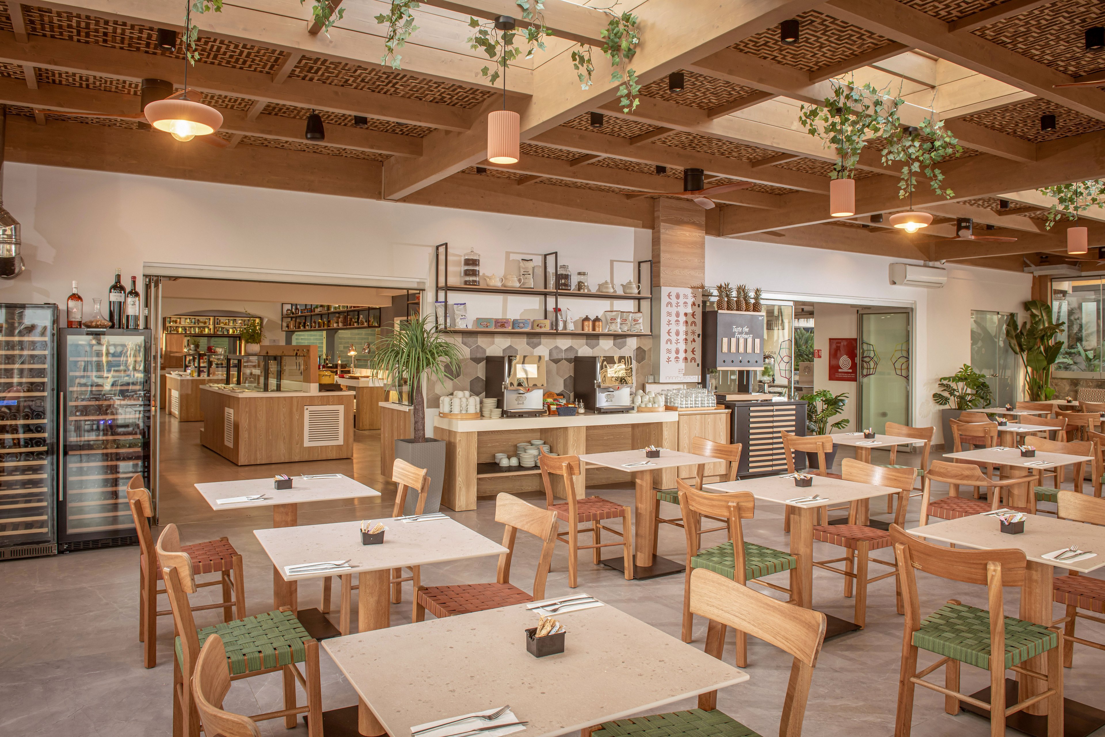 Spacious café interior featuring light wooden tables and chairs arranged neatly in an open dining area. The setting includes a service counter with coffee equipment, shelves of jars and containers, and a refrigerated display case on the side. Warm neutral tones, green chair cushions, and hanging plants from a patterned wooden ceiling create a natural, inviting atmosphere. The space appears clean, organized, and ready to welcome guests for casual dining or coffee.
