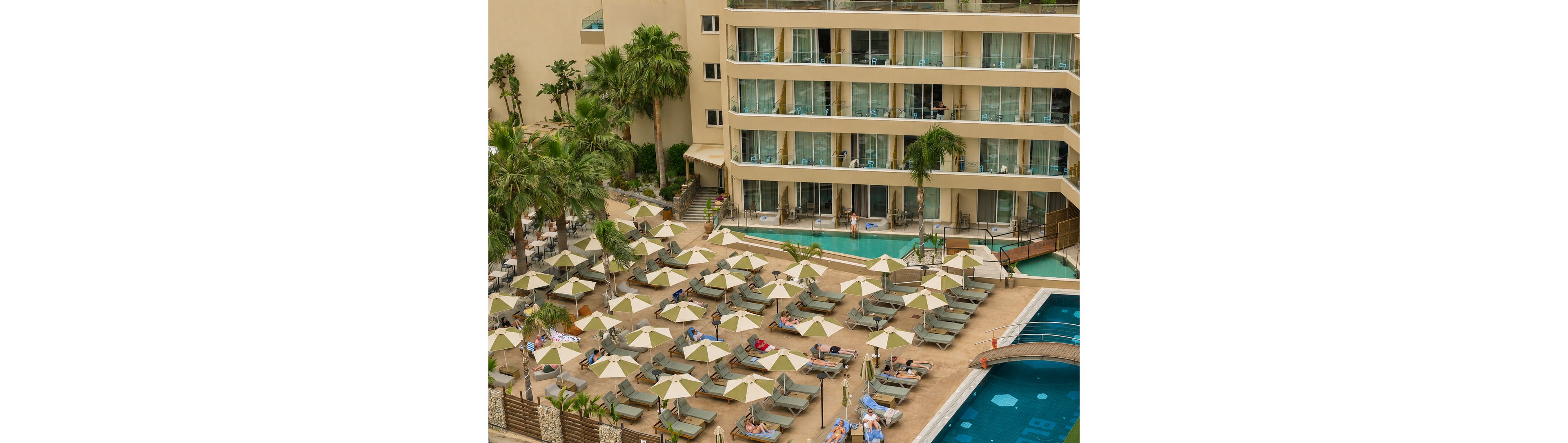 A large hotel resort features multiple stories of balconies overlooking a spacious poolside terrace. Numerous sun loungers with beige umbrellas are arranged in neat rows around the swimming pool. Guests relax on the loungers and near the water, creating a lively vacation atmosphere. Tall palm trees and landscaped areas add greenery and a tropical feel to the scene.