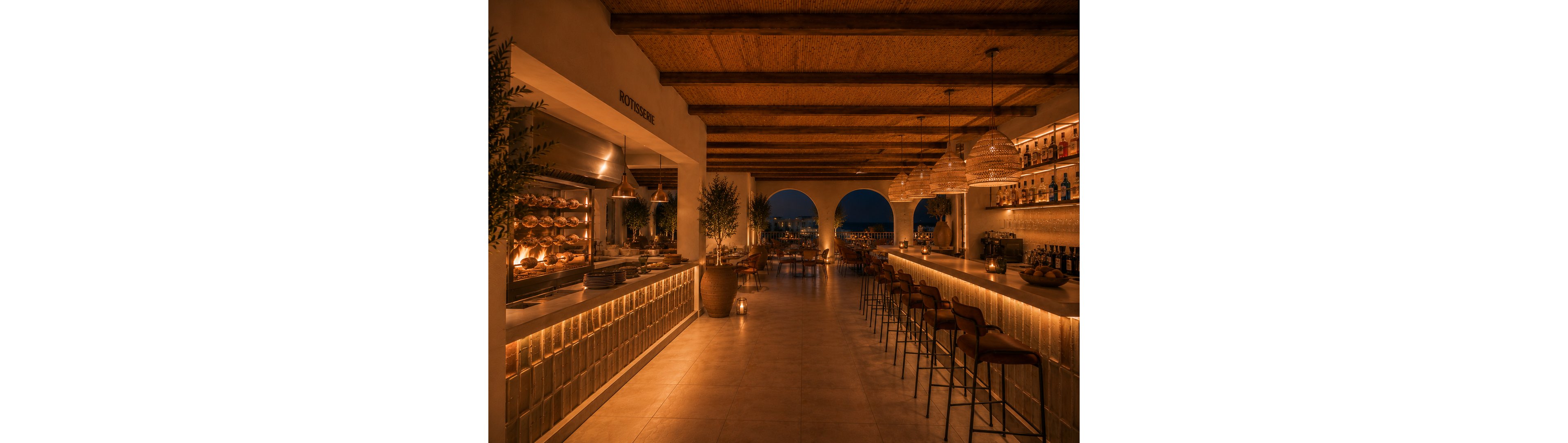 An elegant open-air bar and restaurant is softly lit with warm ambient lighting at dusk. The long counter with high stools faces a rotisserie kitchen on one side and a back bar with bottles and glassware on the other. Arched openings reveal a distant cityscape and evening sky, while candles and pendant lamps create a cozy, intimate atmosphere. Natural materials, potted plants, and woven ceiling details contribute to a relaxed Mediterranean-inspired setting.