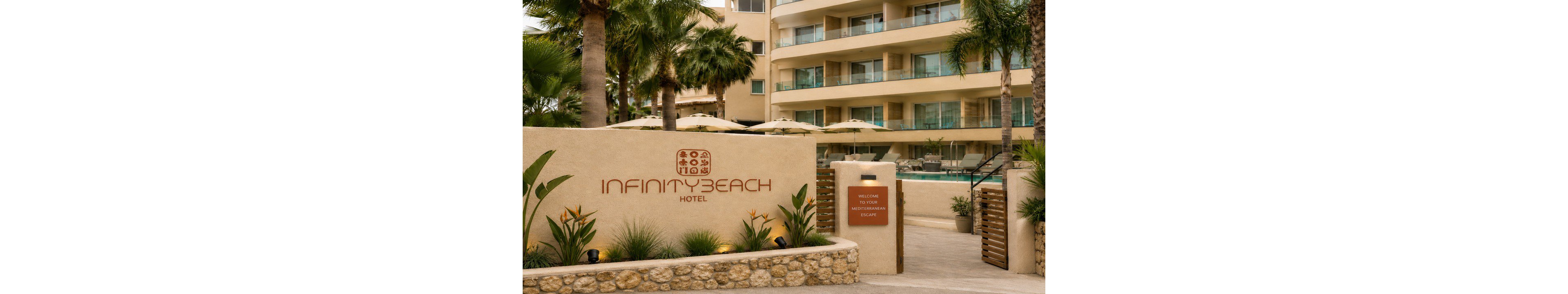 Exterior view of the Infinity Beach Hotel entrance with a stone wall and landscaped plants in the foreground. A sign welcomes guests to the Mediterranean lounge, while palm trees and umbrellas frame the pool area behind the wall. The multi-story hotel building with balconies overlooks the pool, creating a relaxed resort atmosphere. Warm neutral tones and tropical greenery suggest a coastal vacation setting.