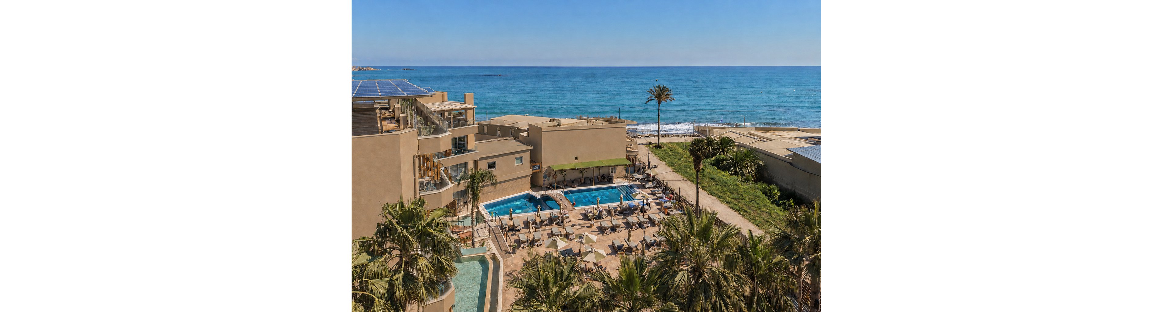 A coastal resort overlooks a bright blue sea with waves gently reaching the shore. Multiple outdoor swimming pools are surrounded by sun loungers and umbrellas, where guests relax under clear skies. Palm trees and greenery frame the terrace areas, adding a tropical feel to the modern buildings. Solar panels are visible on the rooftops, suggesting an eco-conscious design.