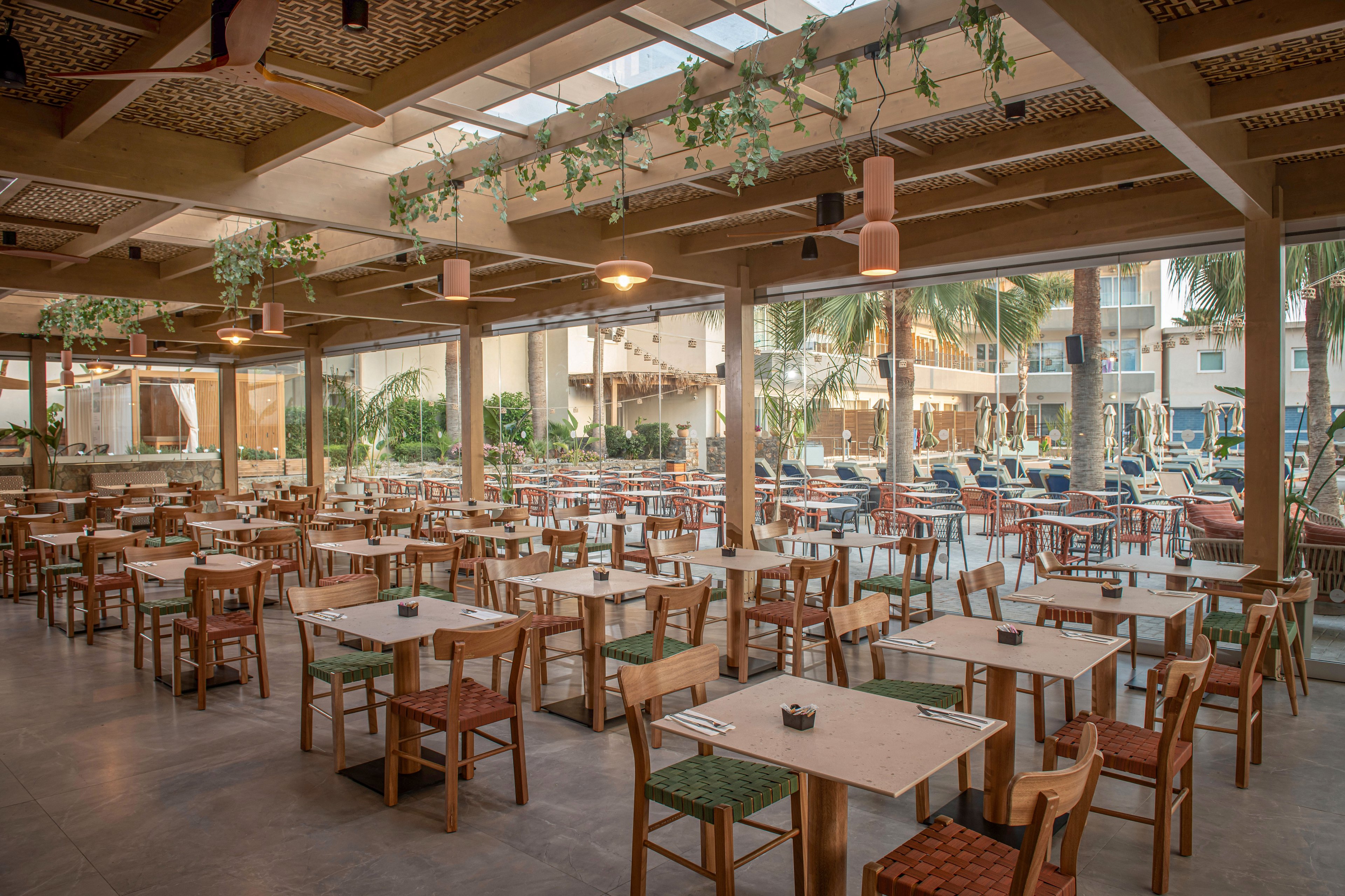 Spacious open-air restaurant with rows of wooden tables and chairs is shown in a resort setting. Large floor-to-ceiling windows reveal an outdoor pool area with sun loungers and palm trees. Warm pendant lighting, ceiling fans, and hanging greenery create a relaxed, tropical atmosphere. The space appears ready for guests, with neatly arranged place settings on each table.