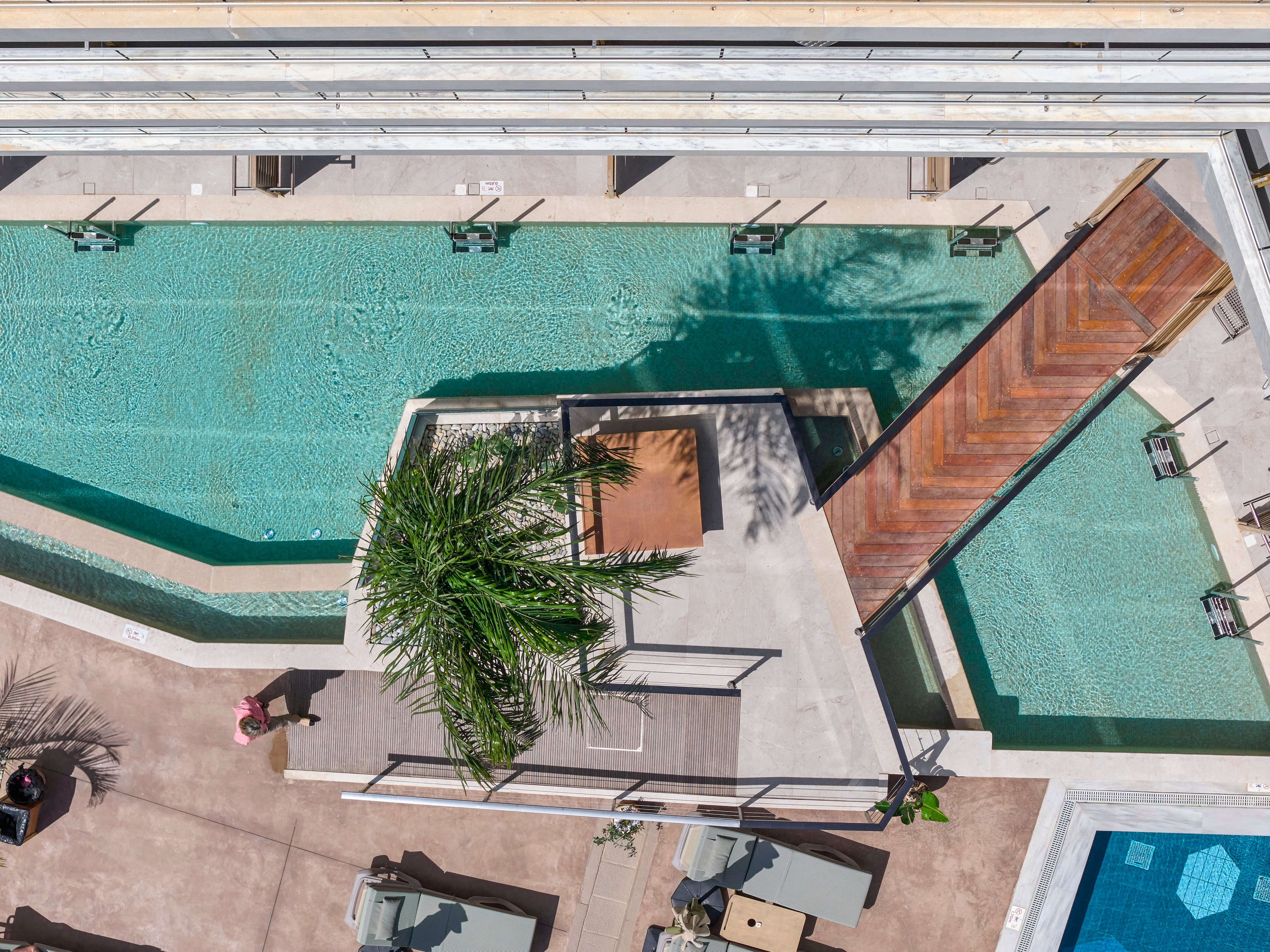 A contemporary outdoor swimming pool complex is seen from above, featuring multiple rectangular pools and a central deck area. A wooden walkway crosses one of the pools, connecting different lounging zones. Palm trees and sun loungers surround the water, suggesting a relaxed vacation setting. Clear turquoise water and geometric design elements create a clean, modern aesthetic.