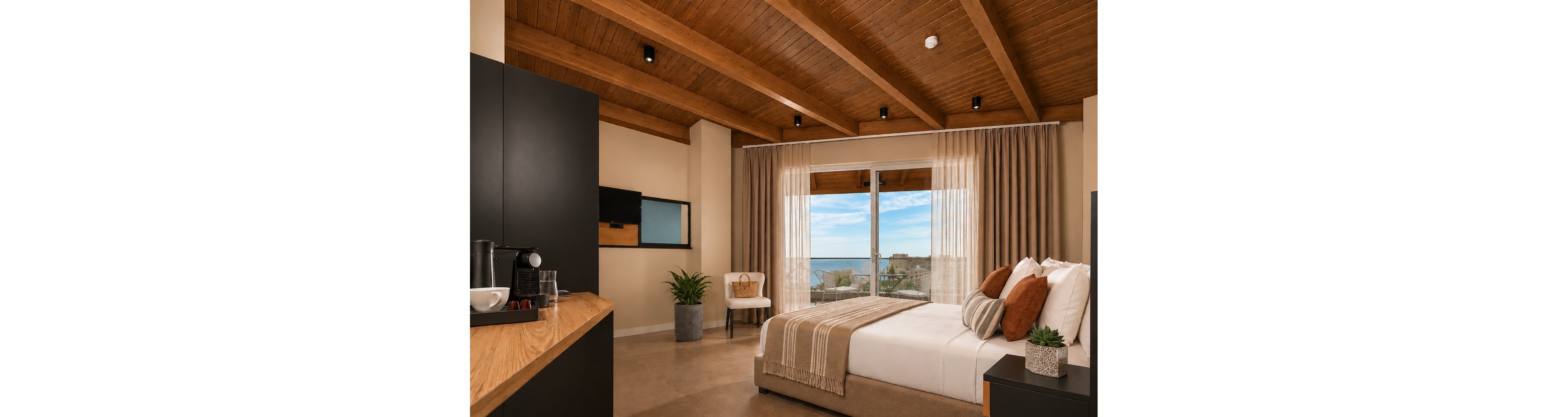 Contemporary hotel room features a neatly made double bed with neutral pillows and a textured throw blanket. Large glass doors with sheer curtains open to a balcony overlooking the ocean and distant buildings under a blue sky. The space includes a wooden ceiling with recessed lights, a small seating area with a chair and plant, and a sleek black cabinet with a coffee station. Overall, the room feels warm, inviting, and designed for comfortable seaside relaxation.