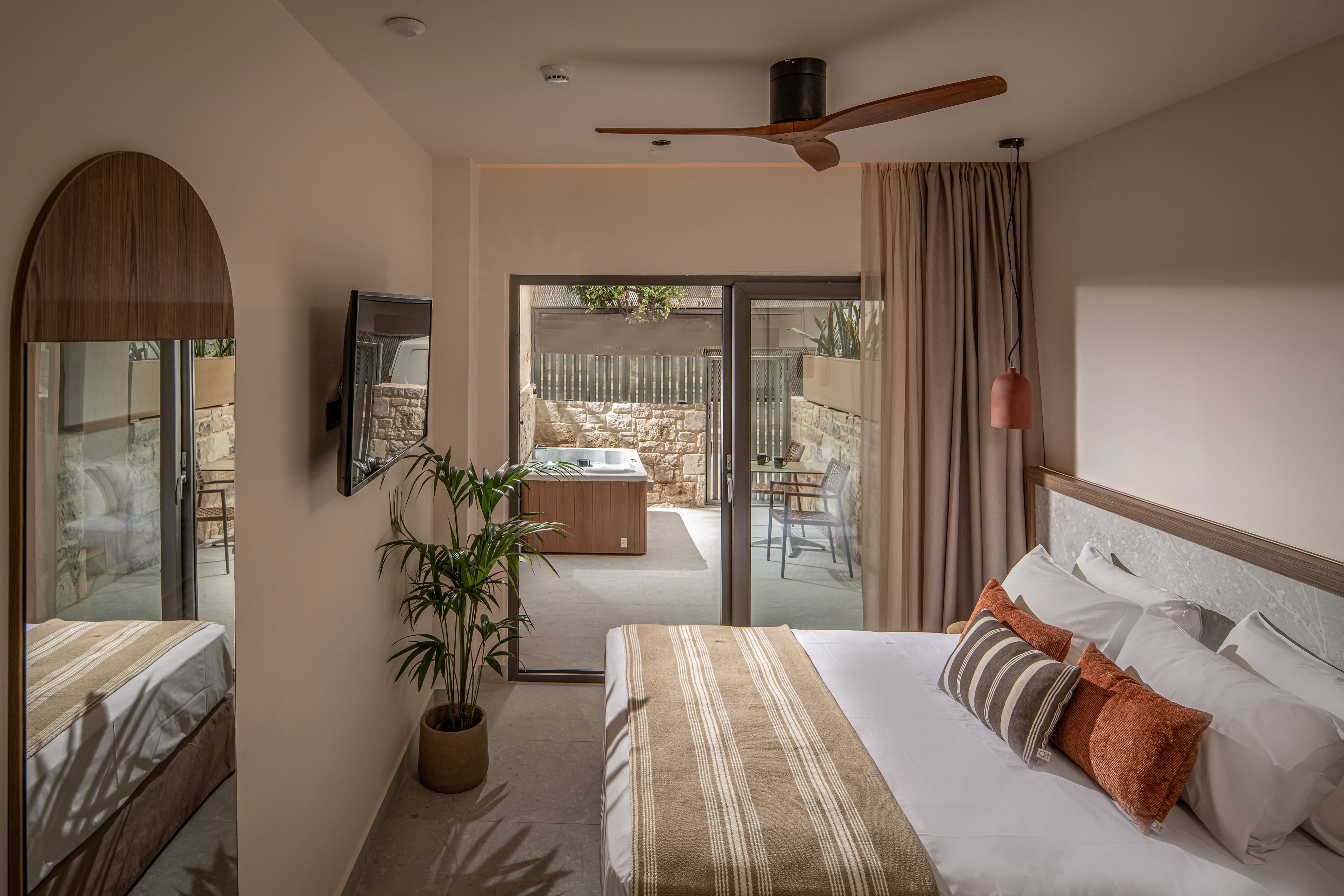 A contemporary hotel bedroom features a neatly made double bed with striped bedding and decorative cushions. The room includes a wall-mounted television, a wooden console, and a potted plant adding greenery. Large sliding glass doors open to a private terrace with outdoor seating and a hot tub surrounded by stone walls. Warm neutral tones and soft lighting create a cozy, inviting atmosphere.