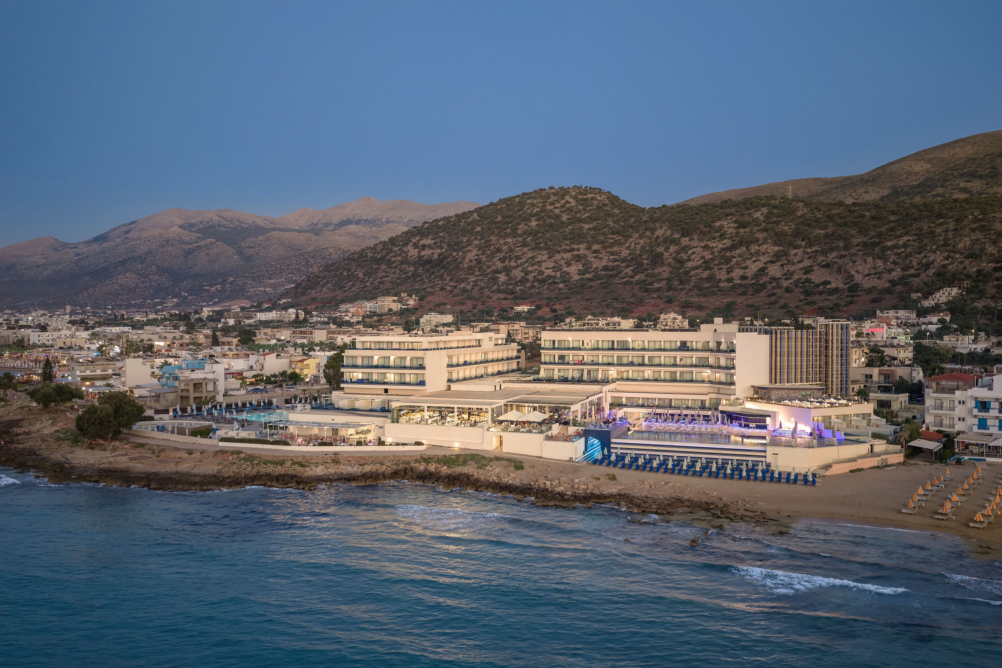 A large seaside resort complex stretches along a calm shoreline at dusk. Multiple hotel buildings face the water, with rows of beach chairs and umbrellas arranged on the sand. Soft artificial lighting highlights parts of the property, contrasting with the fading natural light in the sky. Hills and scattered buildings rise in the background, framing the coastal setting and emphasizing the tranquil vacation atmosphere.