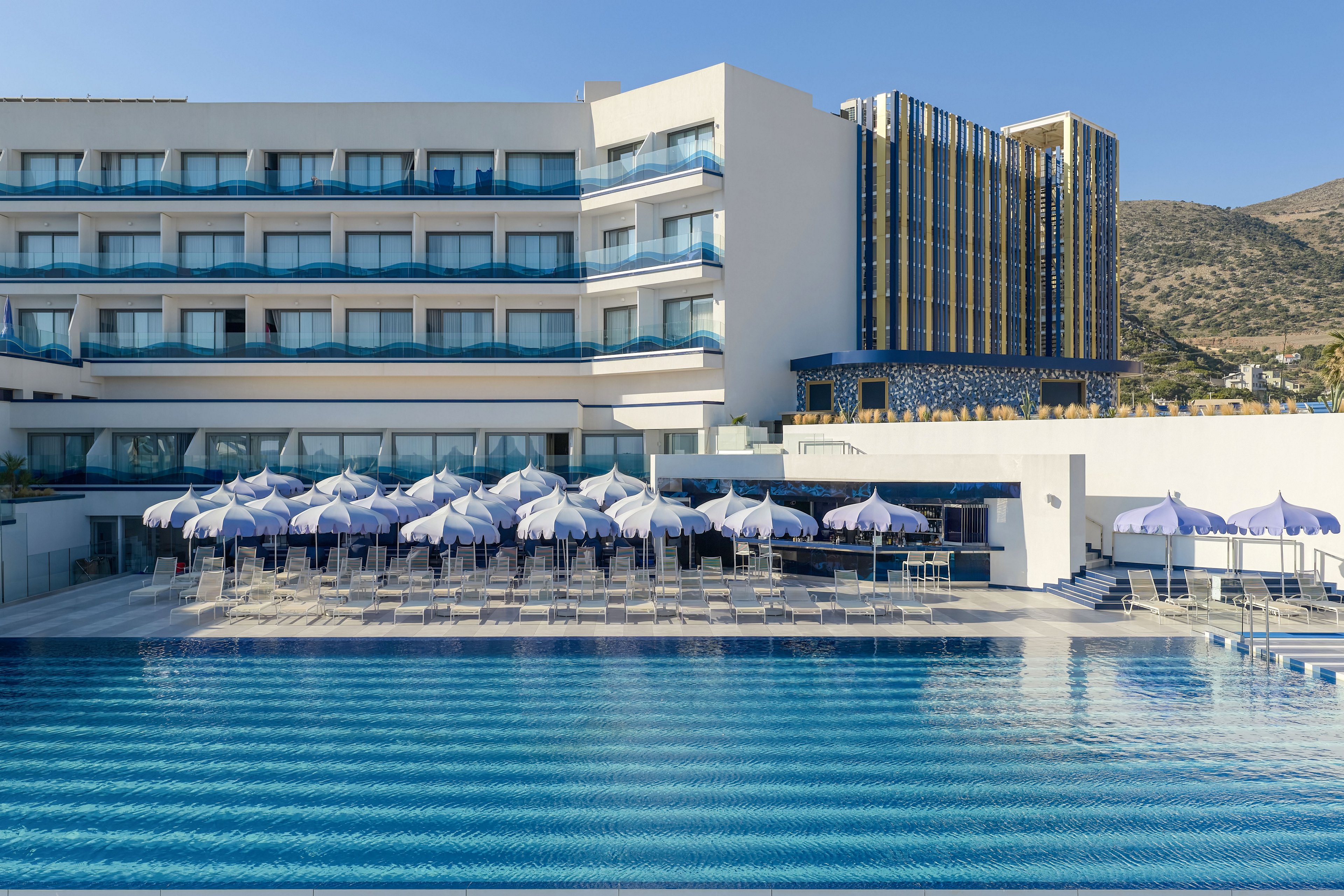 A contemporary multi-story hotel stands beside a large outdoor swimming pool under a clear blue sky. Rows of white and blue sun umbrellas and sun loungers line the poolside terrace, suggesting a resort setting for relaxation and leisure. The building features balconies with glass railings and blue accents, along with a taller tower section and hillside landscape in the background. The calm water and clean architectural lines create a bright, inviting vacation atmosphere.