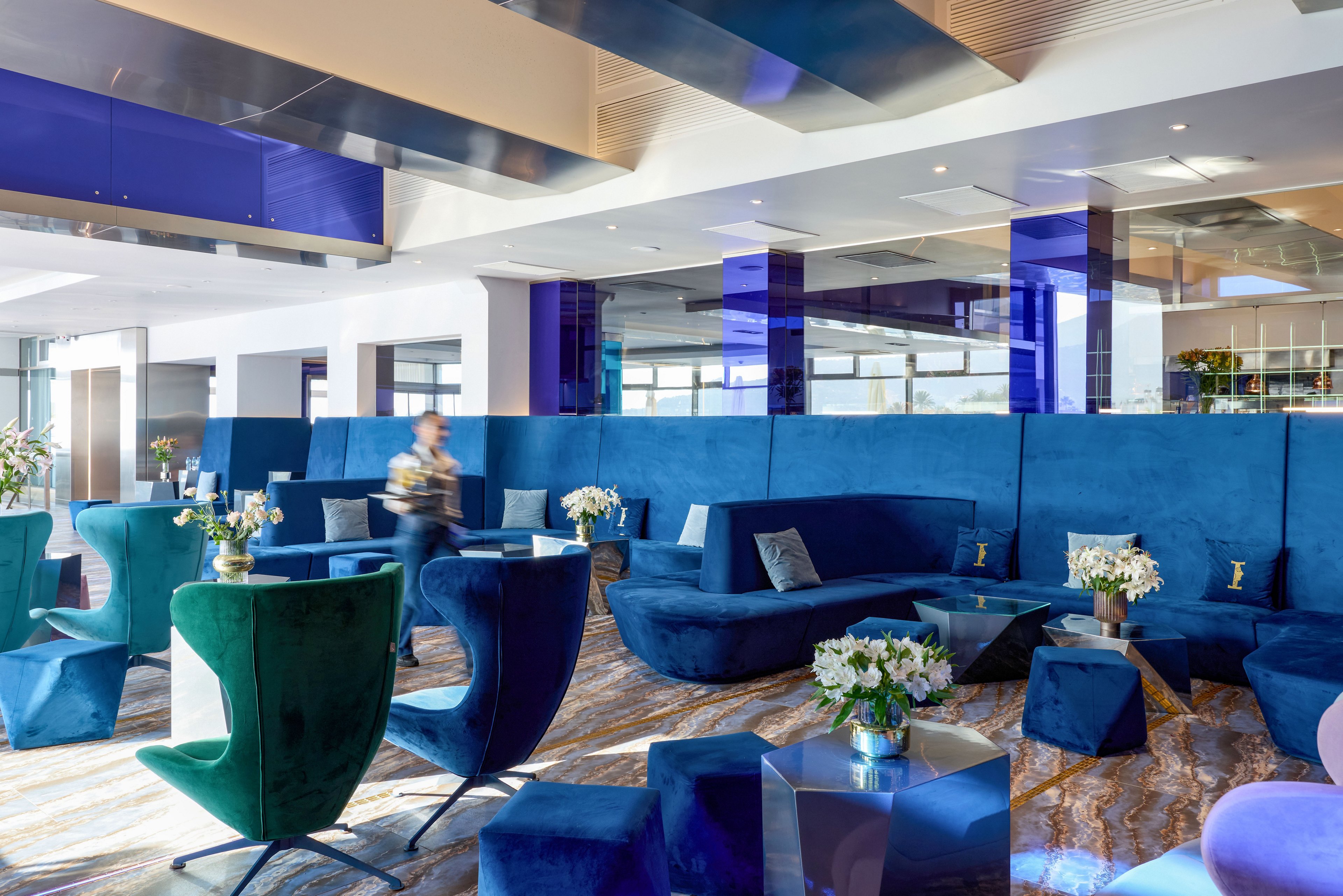A contemporary lounge area features bold blue upholstered sofas and sculptural chairs arranged around low metallic tables. The setting appears to be a bright indoor public space with large windows and reflective ceiling panels. A blurred person walks through the scene, adding a sense of motion and activity. Fresh flower arrangements and geometric furniture pieces contribute to a stylish, upscale atmosphere.