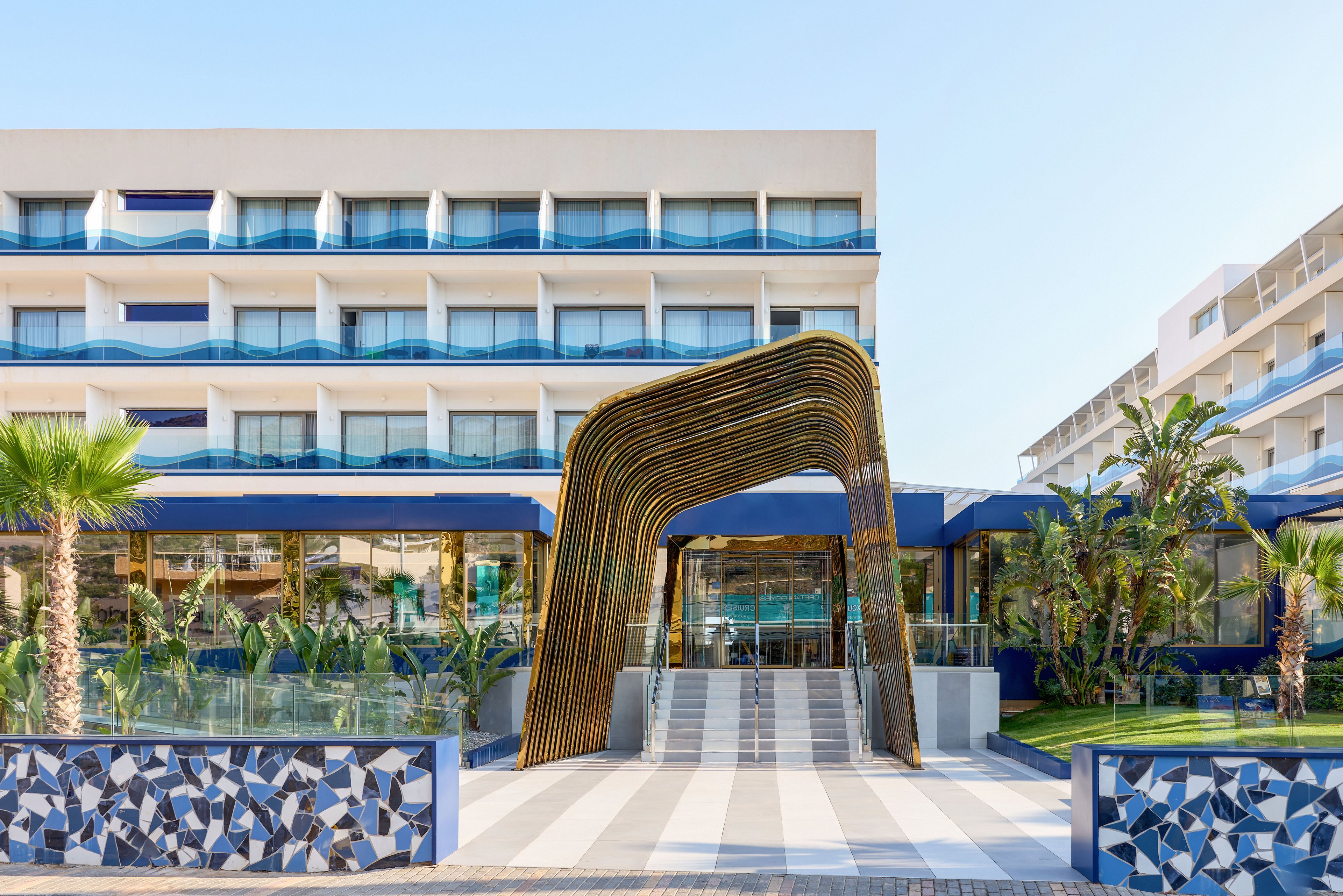 A contemporary multi-story hotel features a striking sculptural archway at its main entrance. The building has rows of balconies with glass railings and blue accents, suggesting a coastal or resort setting. Palm trees and lush greenery line the front, complemented by geometric patterned tiles along the low wall. Bright daylight and clear skies create a clean, inviting atmosphere with a modern architectural style.