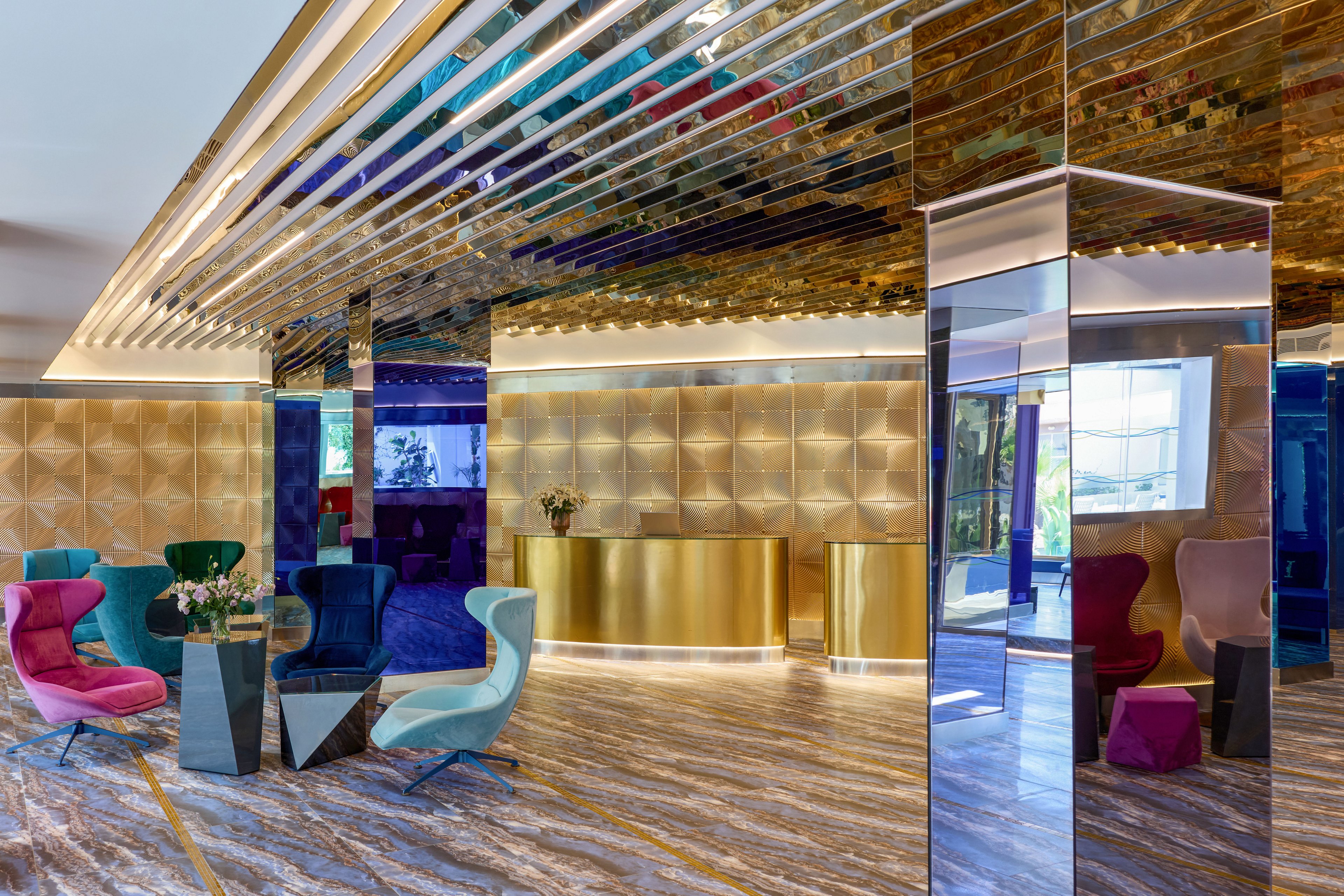 A contemporary lobby features a golden reception desk set against a textured metallic wall. Colorful, sculptural lounge chairs surround sleek black side tables with flower arrangements. The ceiling and columns are clad in reflective metallic panels, creating a luxurious and futuristic atmosphere. Large windows bring in natural light, highlighting the polished patterned floor and vibrant interior design.