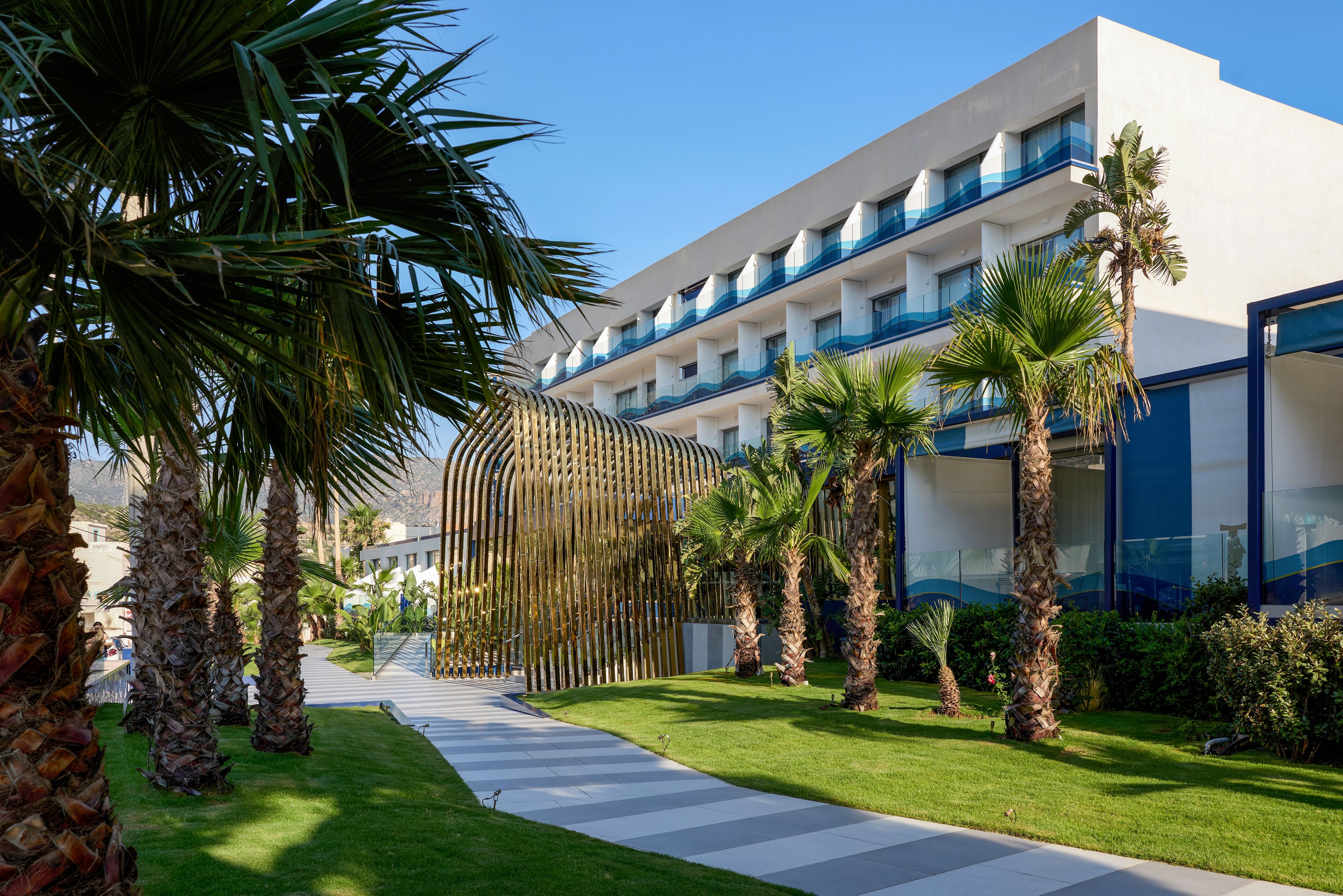 A contemporary multi-story hotel building is shown with balconies facing a landscaped garden. A curved metallic architectural structure stands beside a striped walkway leading through the green lawn. Tall palm trees line the path, adding a tropical atmosphere to the scene. Clear blue sky and distant hills suggest a sunny coastal or resort location.