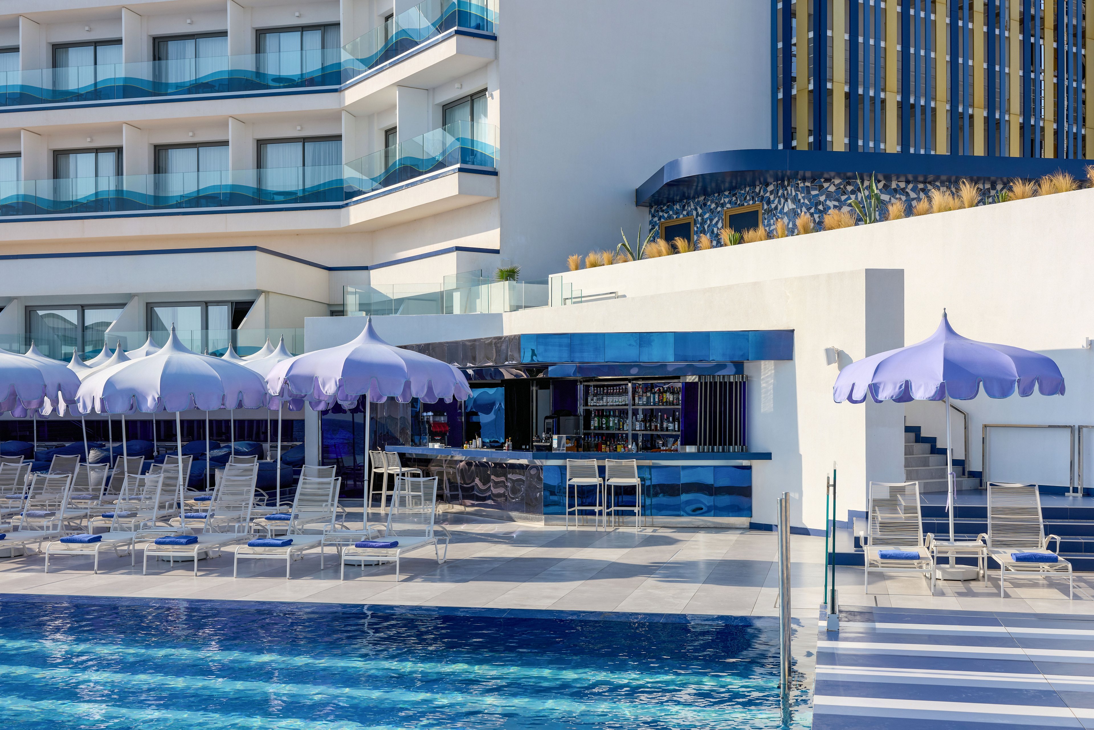 A contemporary hotel pool area features a sleek bar surrounded by lounge chairs and pastel umbrellas. The setting is outdoors, with the pool in the foreground and multi-story balconies overlooking the scene. Blue and white tones dominate the architecture and pool tiles, creating a clean, coastal atmosphere. The area appears calm and ready for guests, suggesting a relaxing vacation or resort experience.