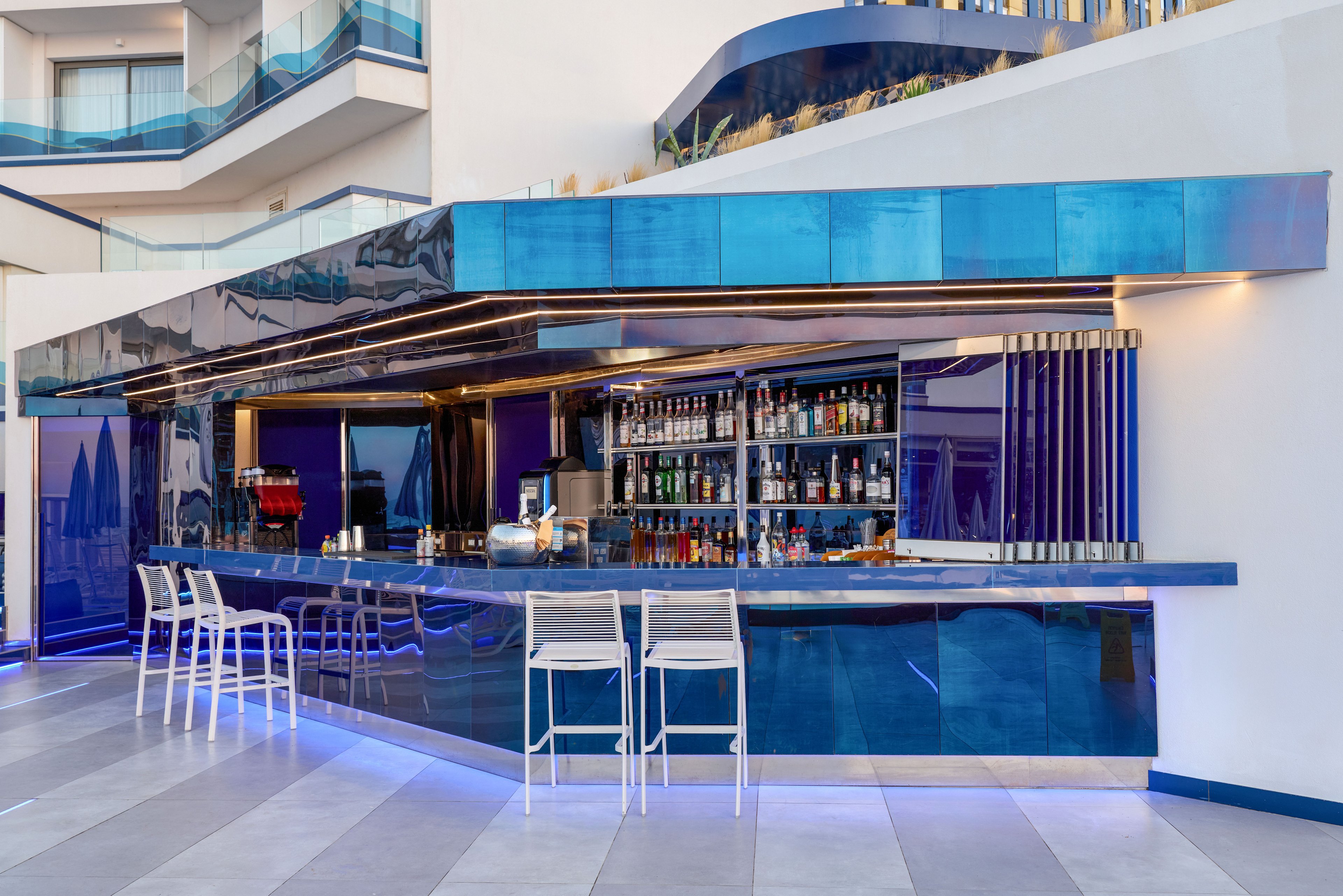 A sleek contemporary bar with a glossy blue counter and metallic accents stands in an open hotel terrace area. Shelves behind the bar are stocked with various bottles and glassware, creating a vibrant focal point. Several white bar stools line the counter, while architectural details and balconies of the hotel rise in the background. Cool blue lighting and reflective surfaces give the space a stylish, upscale atmosphere.