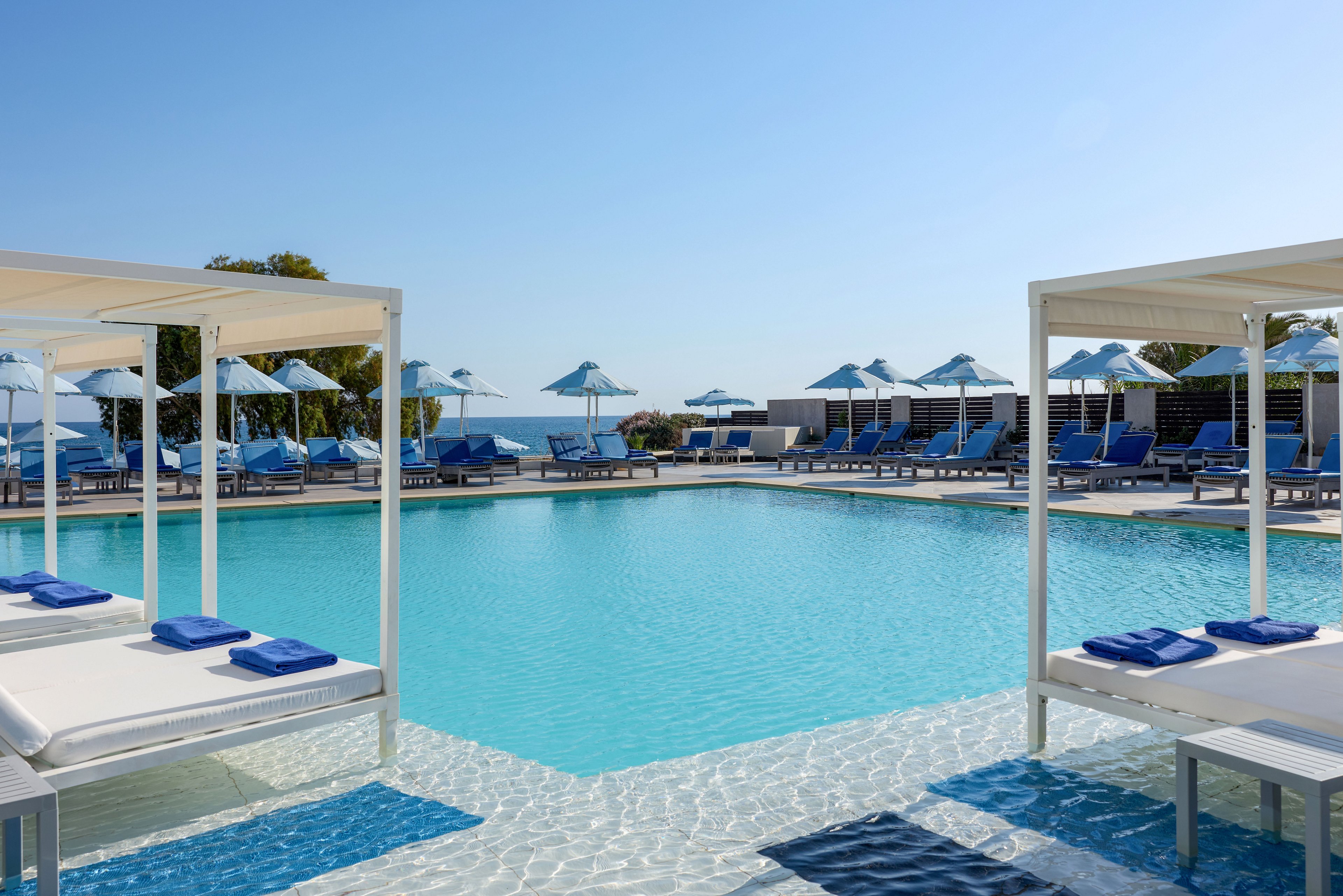 A tranquil resort swimming pool is surrounded by white cabanas and blue sun loungers. The clear water reflects the bright blue sky, creating a serene vacation atmosphere. Umbrellas and chairs line the far side of the pool, with a glimpse of the sea in the background. The scene suggests a relaxing summer holiday at a coastal destination.