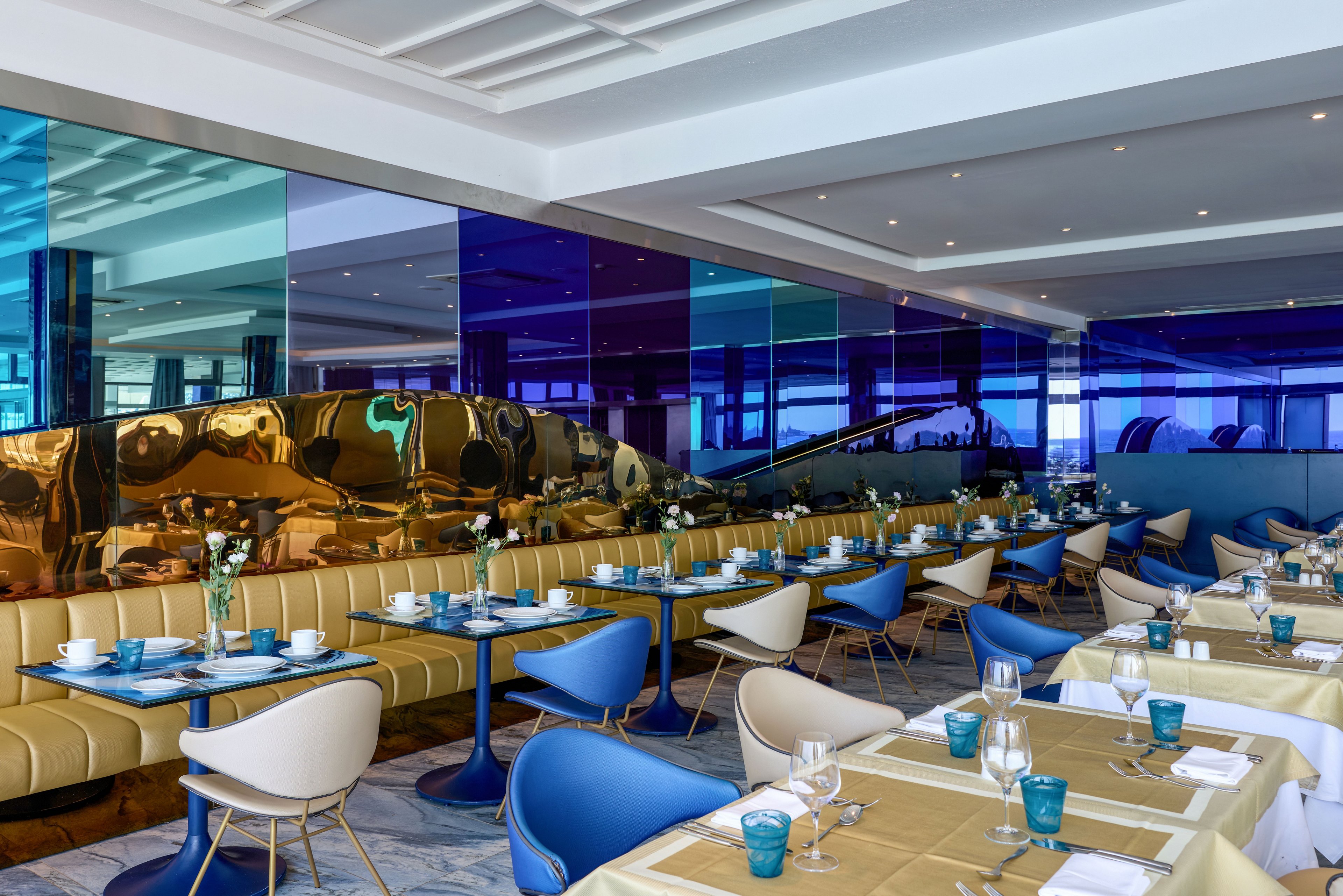 Contemporary restaurant interior featuring neatly arranged tables with beige tablecloths and blue chairs. The setting includes cushioned banquette seating and reflective gold surfaces along the wall. Large panels of blue and purple tinted glass create a vibrant, upscale atmosphere. Tableware, cups, and glasses are set for service, suggesting a prepared dining area in a stylish hotel or resort.