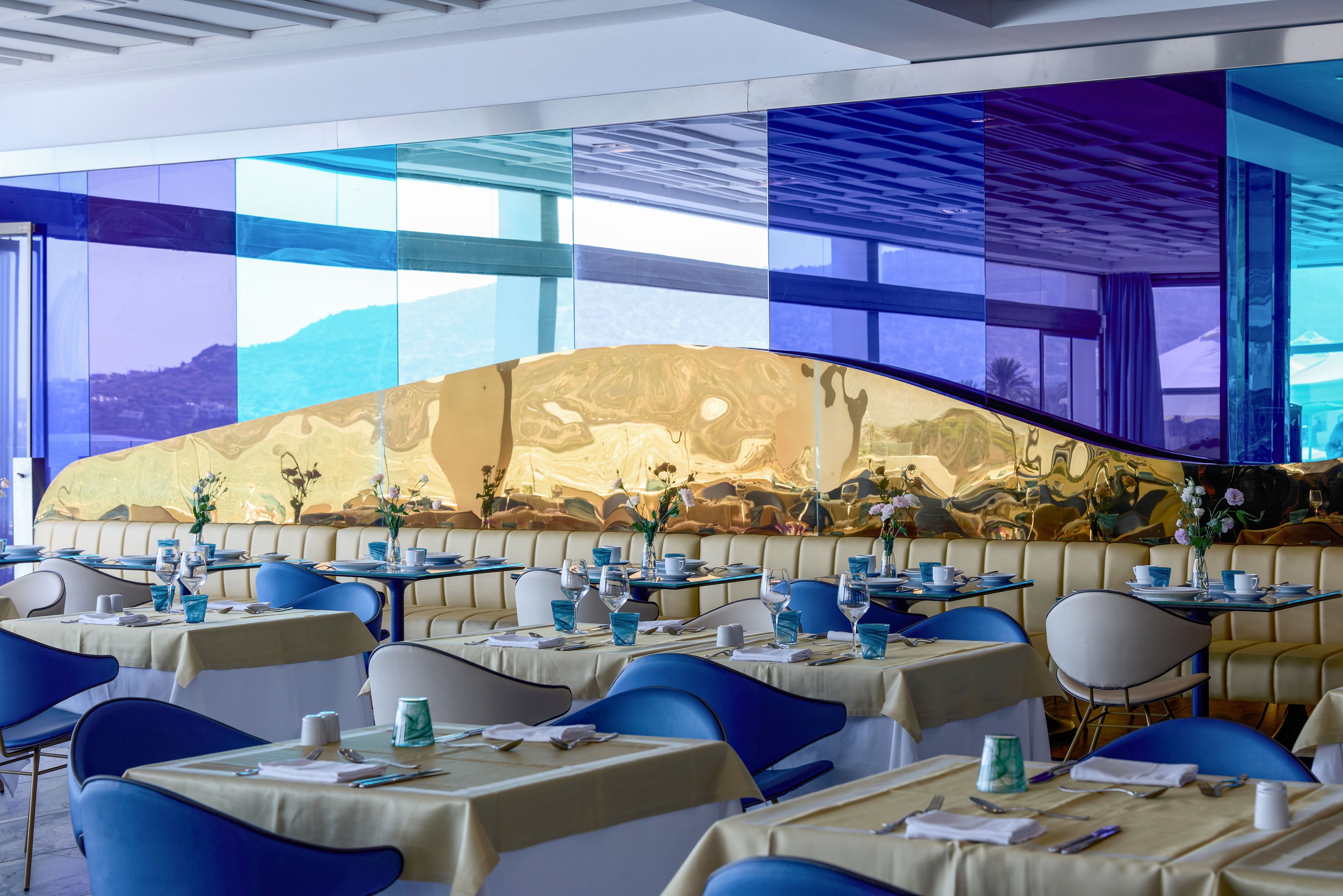 Contemporary restaurant interior featuring neatly arranged tables and blue chairs. A striking wall of blue and purple tinted glass panels frames a view of distant mountains and water. A sculptural golden metallic divider runs along the back of the seating area, adding a reflective accent. Table settings include light yellow tablecloths, turquoise cups, and small floral centerpieces, creating a fresh and stylish atmosphere.