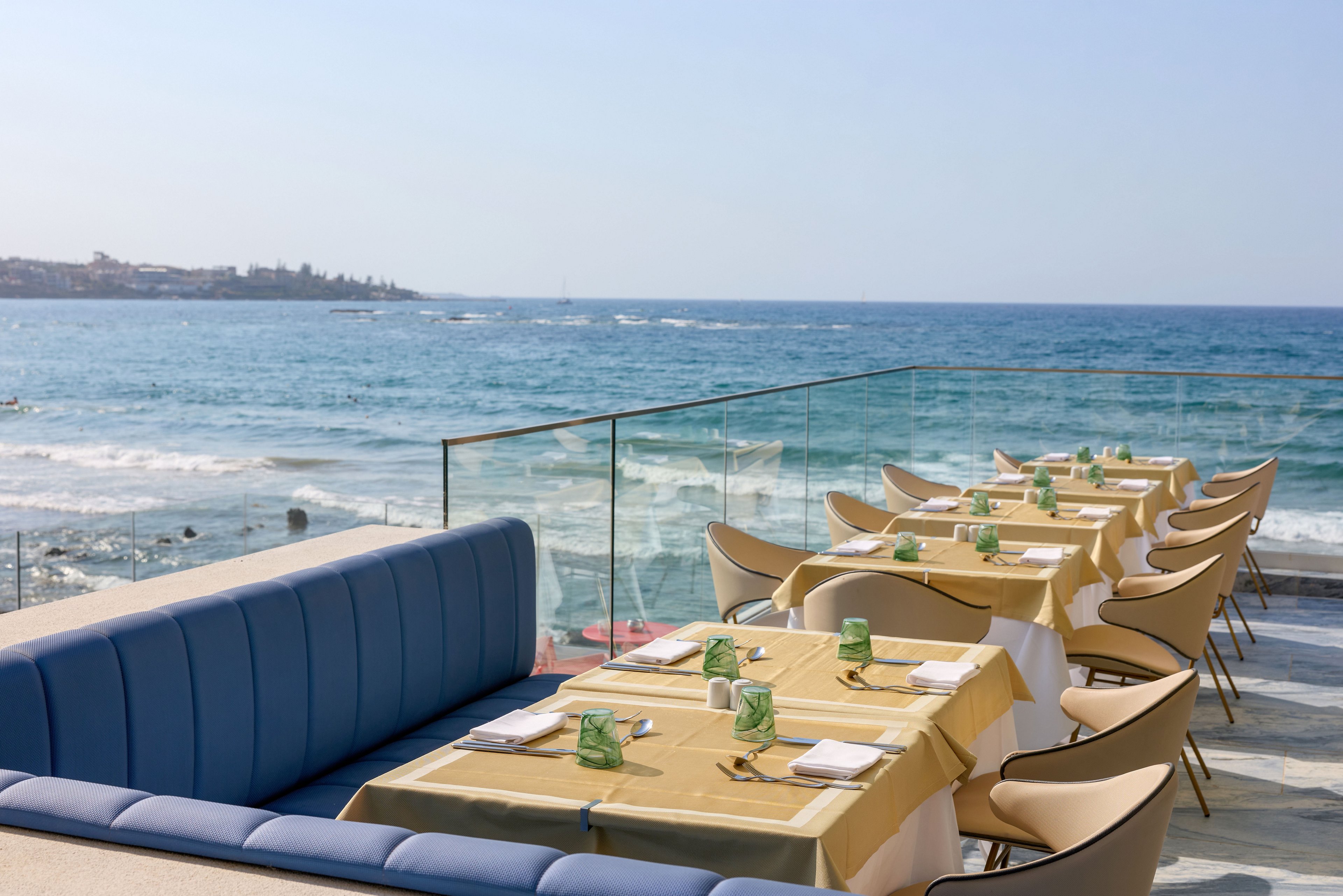 An outdoor restaurant terrace overlooks a calm blue ocean under a clear sky. Tables are neatly arranged with yellow tablecloths, glassware, and place settings beside a long blue cushioned bench. Beige chairs line the tables, and a glass railing separates the dining area from the rocky shoreline below. The distant coastline and gentle waves create a relaxed, coastal atmosphere ideal for al fresco dining.