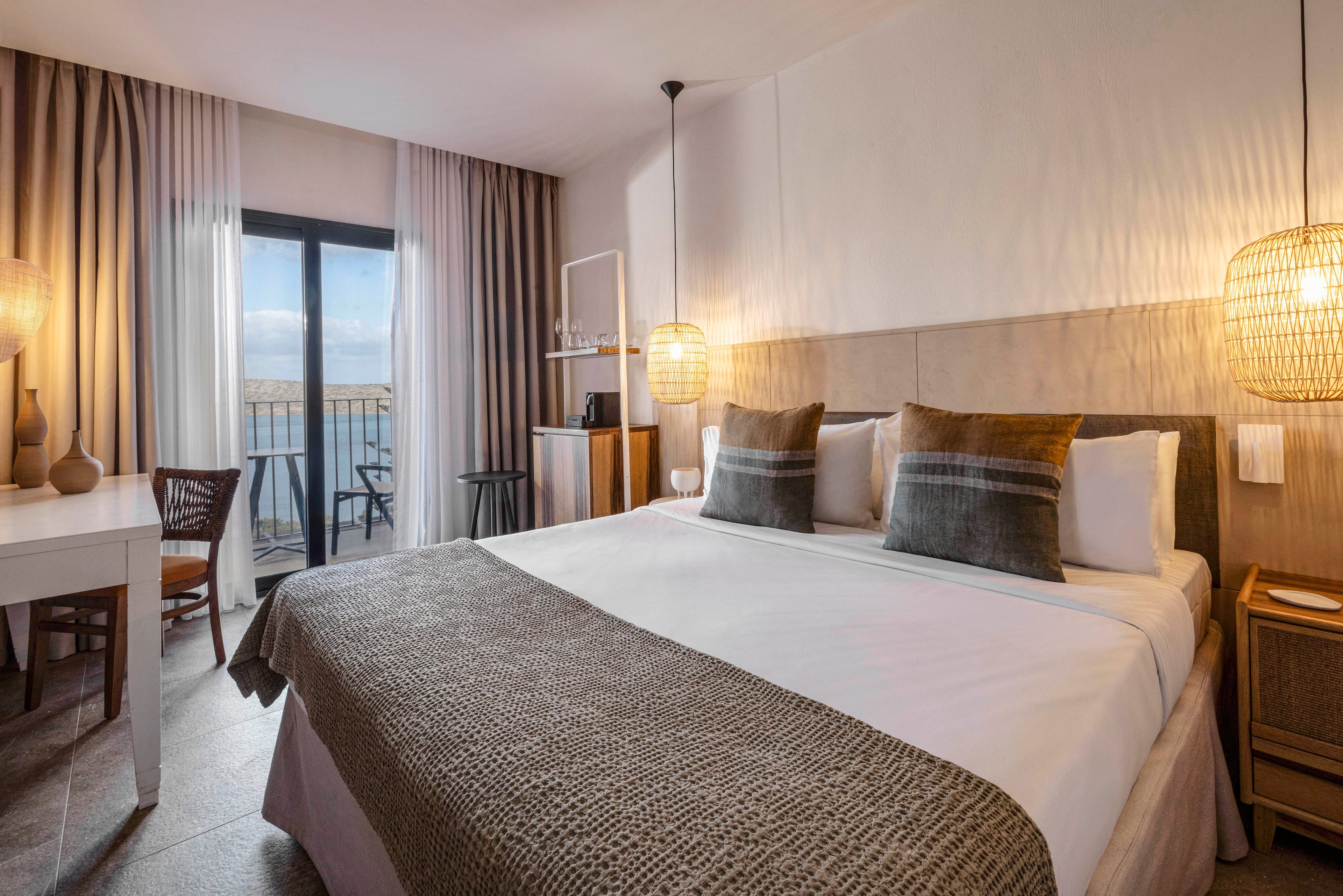 A contemporary hotel room features a large double bed with neutral-toned bedding and accent pillows. The space is illuminated by warm pendant lights and natural daylight from a glass door leading to a balcony with outdoor seating. The room includes a desk, chair, and minimalist decor, creating a calm and inviting atmosphere.