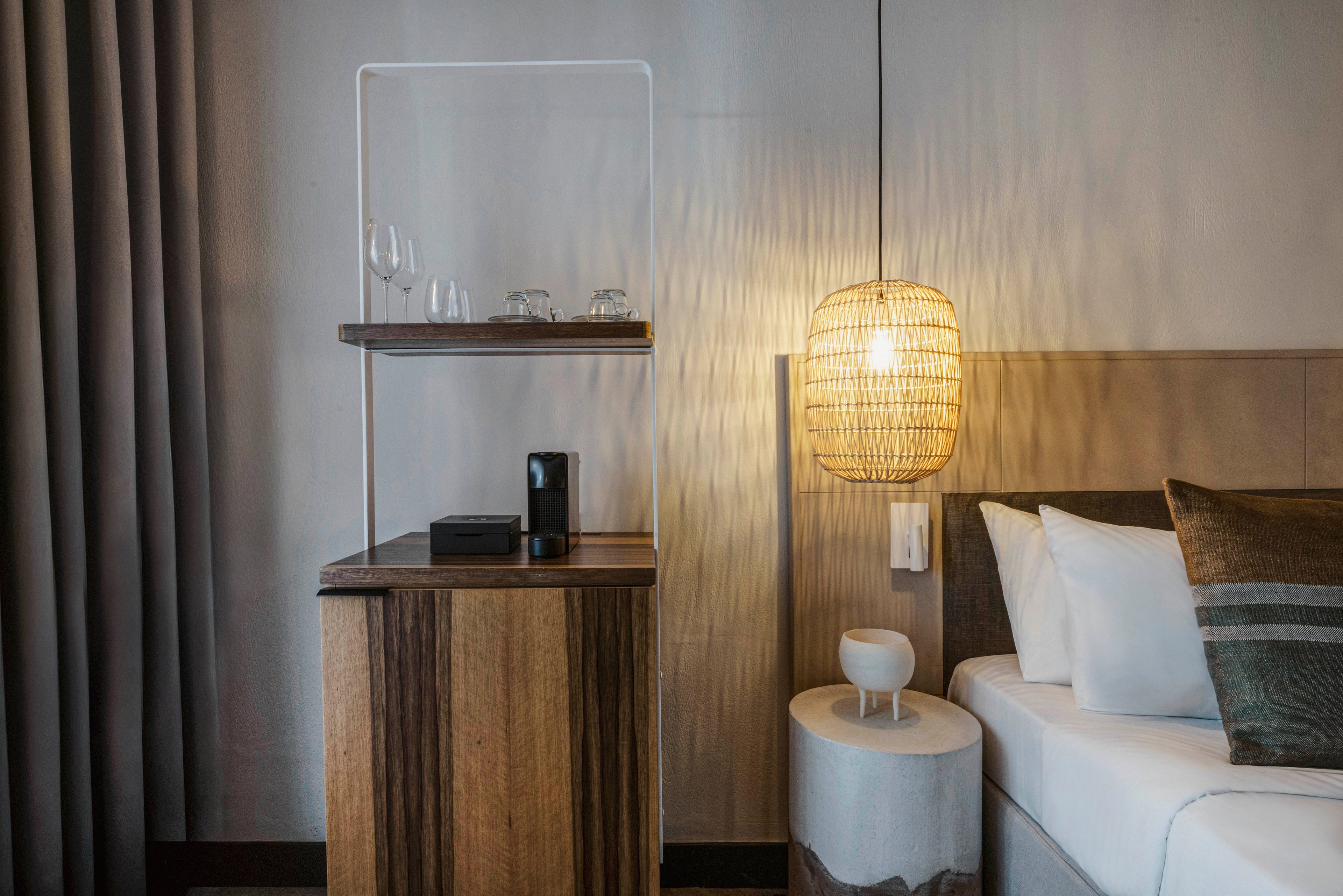 A contemporary hotel room features a wooden nightstand with glassware and a black electronic device on top. A woven pendant lamp hangs above a small round side table next to a neatly made bed with white linens and a striped pillow. The setting is calm and minimalistic, with neutral tones and soft lighting.