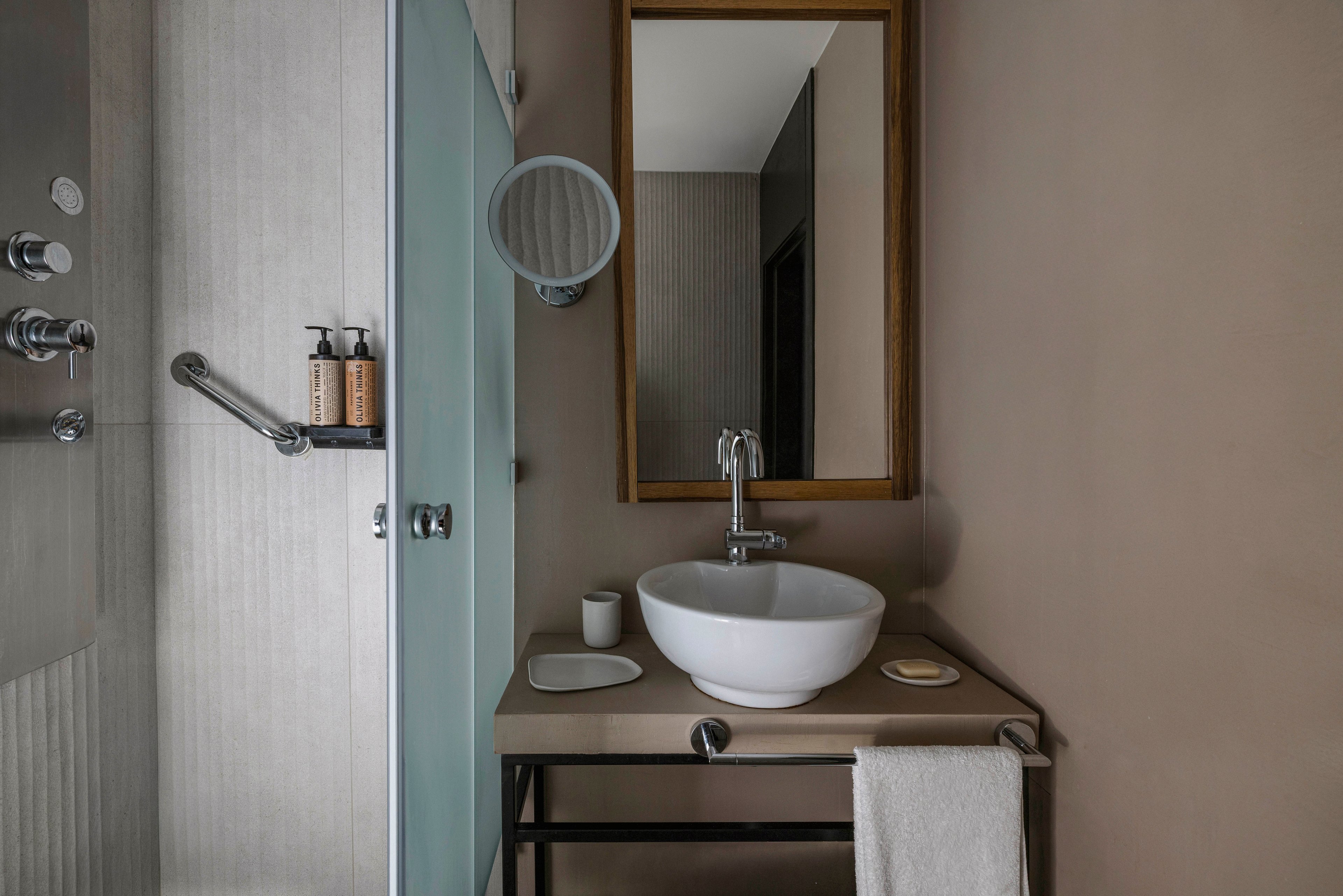 A contemporary bathroom features a white vessel sink atop a beige countertop, complemented by a wall-mounted faucet and a rectangular wooden-framed mirror. The shower area is enclosed with frosted glass and includes stainless steel fixtures and two pump bottles. The neutral color palette and clean lines create a calm, minimalist atmosphere.