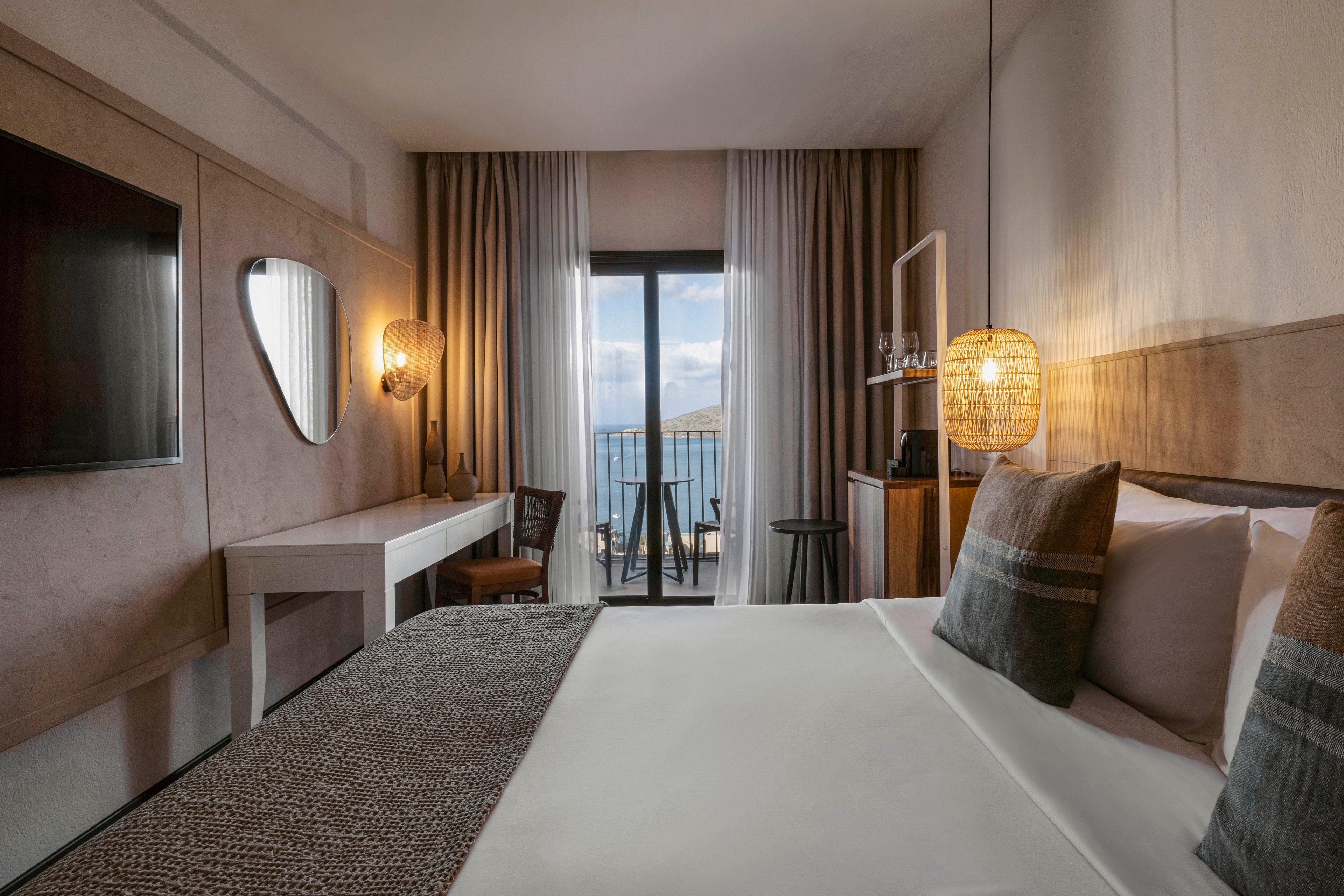 A contemporary hotel room features a large bed with patterned bedding, a wall-mounted television, and a stylish desk area. The room opens to a balcony with two chairs, offering a scenic view of the ocean through glass doors. Warm lighting, neutral tones, and minimalist decor create a calm and inviting atmosphere.