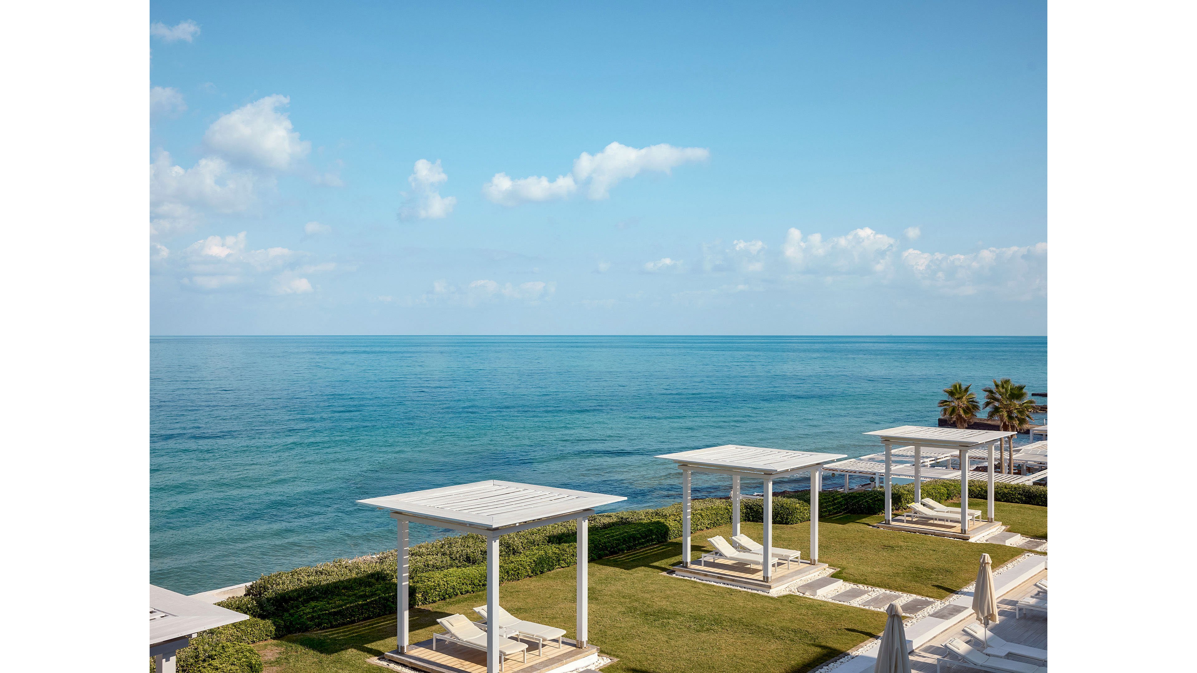 A series of white pergola-style terraces face a calm blue ocean under a clear sky. The scene appears to be part of a modern seaside resort with manicured lawns and hedges. Sun loungers and closed umbrellas line the terraces, suggesting a tranquil vacation setting. Soft clouds and distant horizon enhance the relaxed, coastal atmosphere.