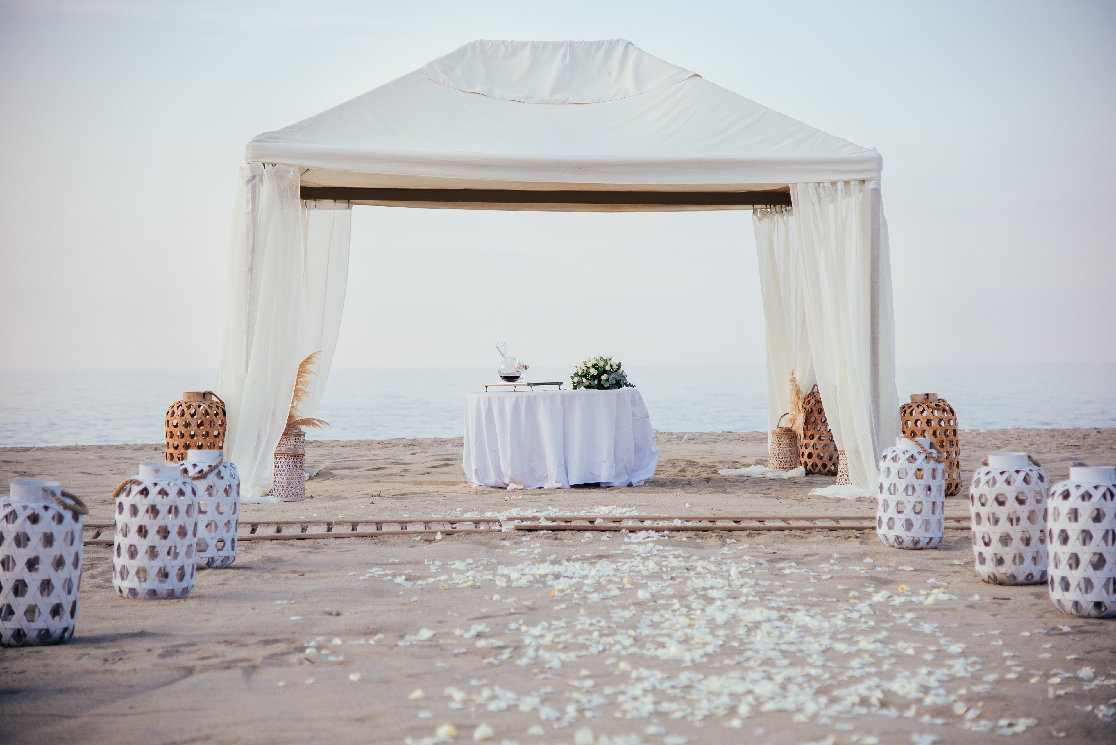 A white gazebo stands on a sandy beach, framing a round table draped in white cloth. The table is decorated with floral arrangements and glassware, suggesting a romantic ceremony or intimate event by the sea. Decorative lanterns and vases line the aisle, with white petals scattered across the sand leading toward the gazebo. The calm ocean and soft sky in the background create a serene, elegant atmosphere.