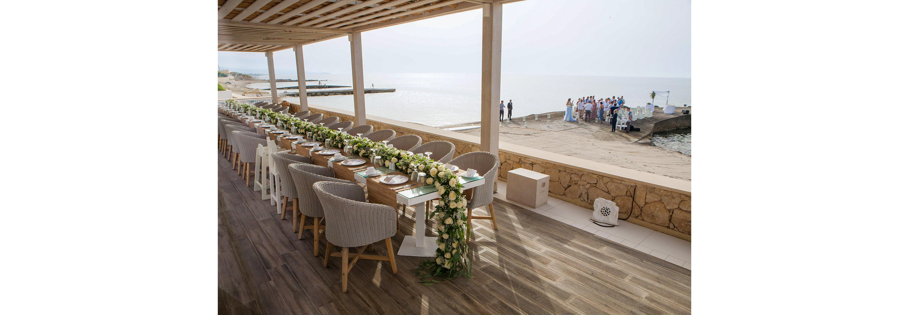A long, elegantly decorated reception table is arranged on a covered terrace overlooking the sea. Soft upholstered chairs line both sides of the table, which is adorned with lush white and green floral garlands and glassware. In the background, a wedding party gathers near an archway on the beach by the water. The scene feels bright and romantic, with natural light and coastal views creating a serene atmosphere.