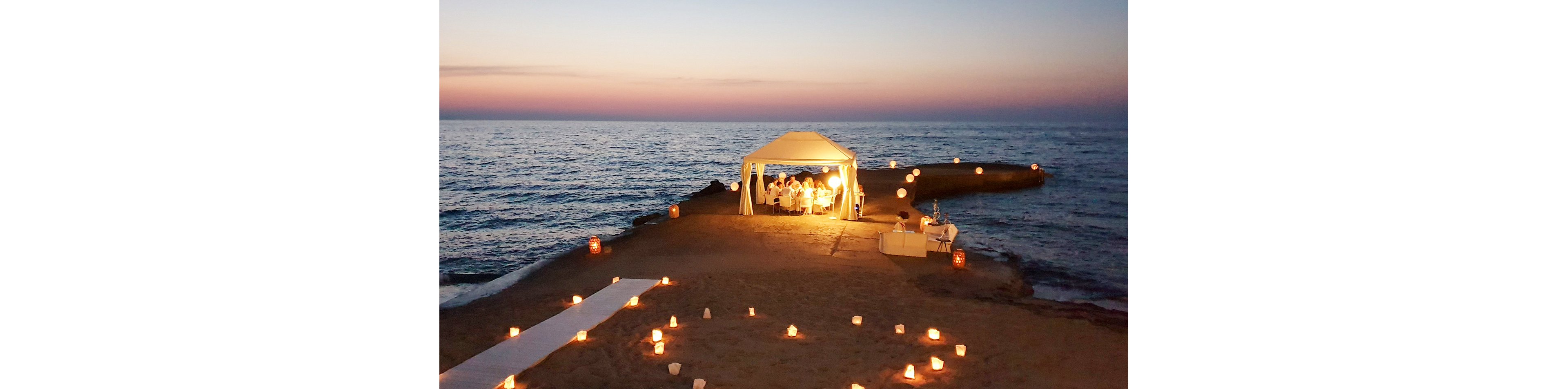 A small group of people gather under a white canopy for an intimate dinner on a pier by the sea at sunset. The sky glows with soft pastel colors as waves gently meet the shoreline. Numerous candles and lanterns illuminate the sand and pathway, creating a warm, romantic atmosphere. A seating area with white furniture adds to the elegant, relaxed beach setting.