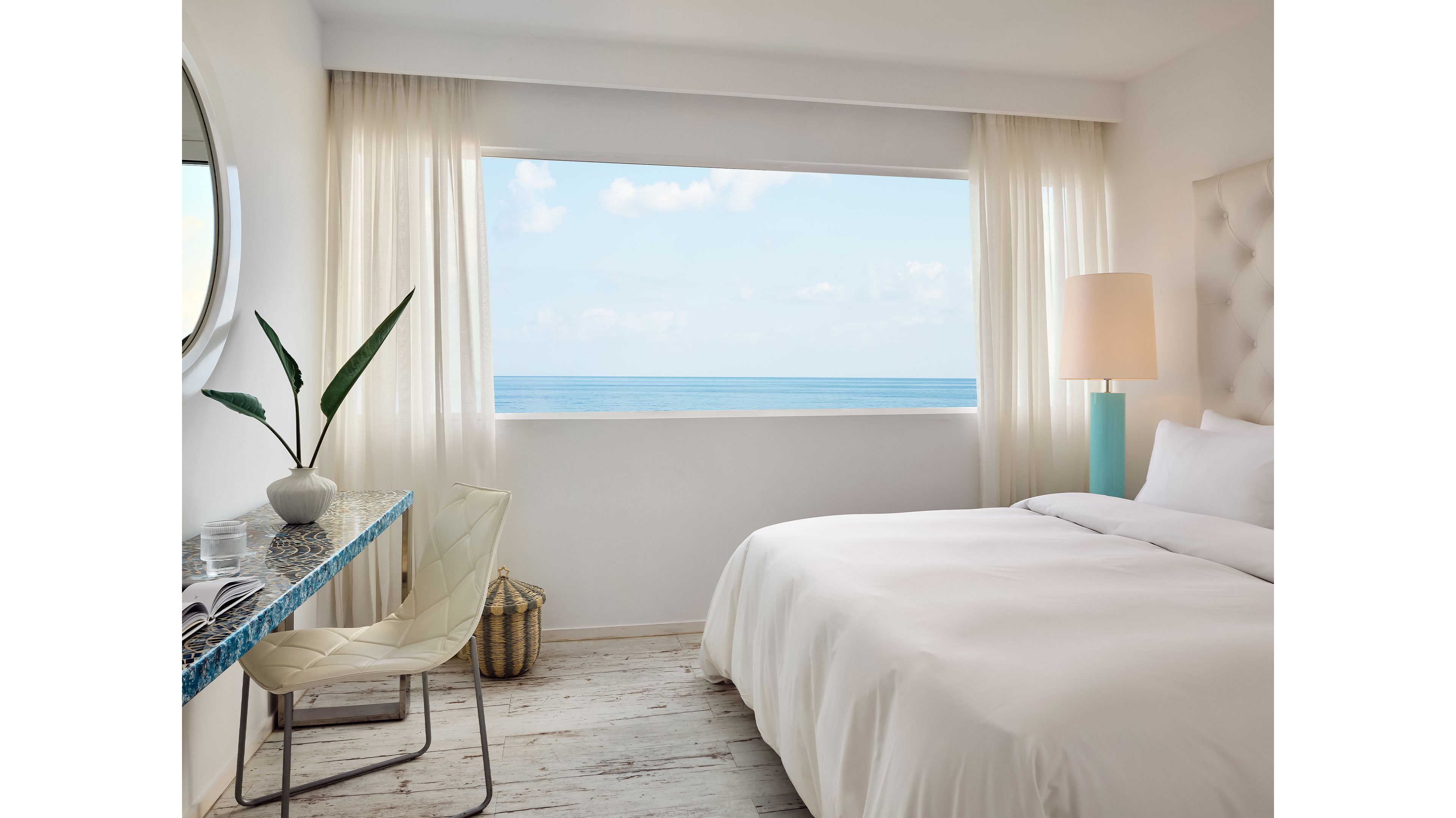 A serene bedroom featuring a large window overlooking the ocean. The room is decorated in a minimalist style with white bedding, light curtains, and a turquoise lamp. A glass desk with a modern chair and a decorative plant add subtle accents to the space.