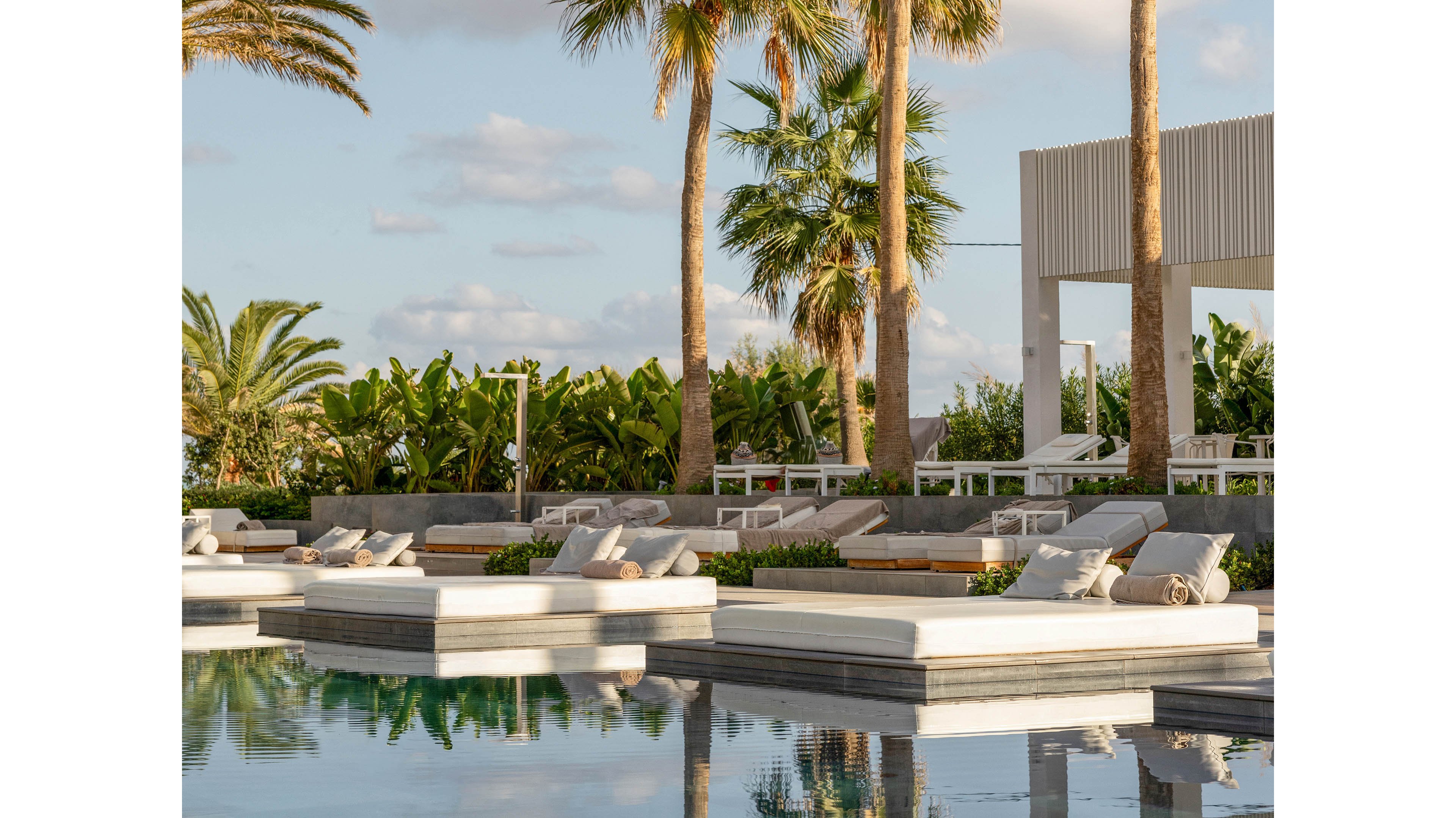 A woman sits on a white sunbed beside a calm swimming pool, reading or using a tablet. The scene is set in a modern outdoor resort area with palm trees and lush tropical plants. Reflections of the loungers and trees shimmer on the still water, enhancing the tranquil atmosphere. Soft afternoon light and clear skies create a relaxed, vacation-like mood.