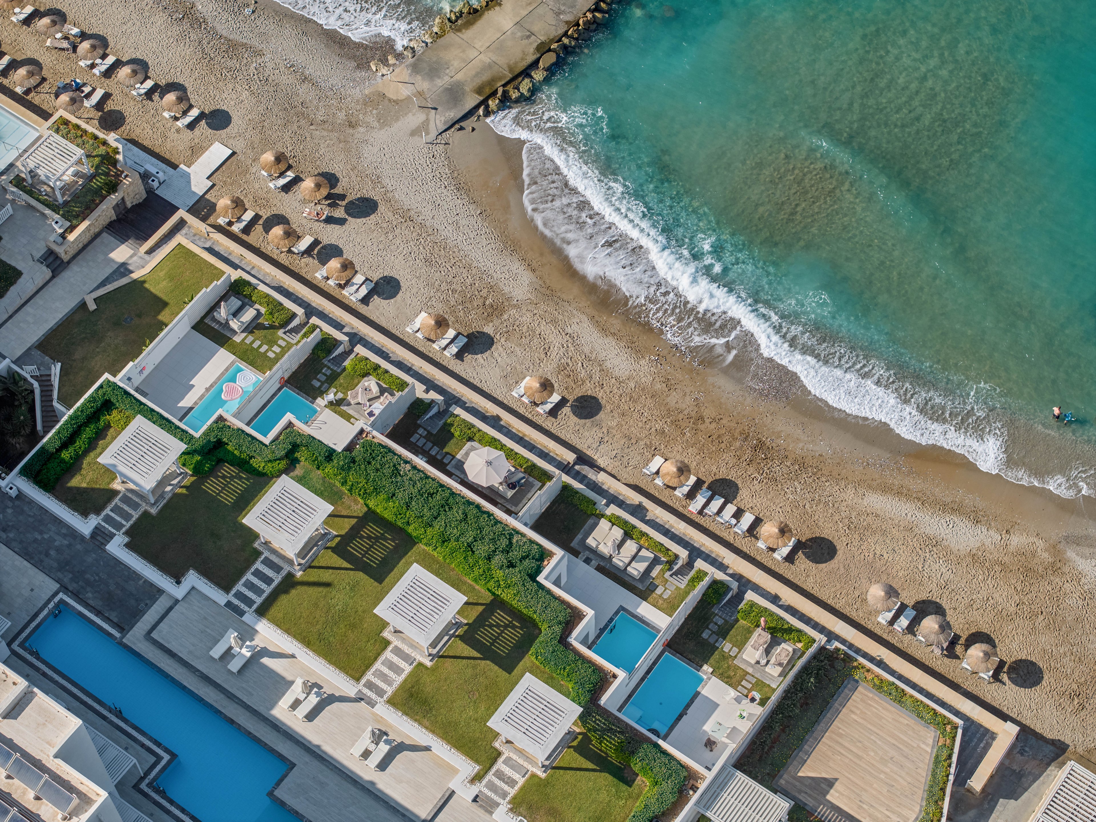 A luxury seaside resort is captured from above, showing modern villas with private pools next to a sandy beach. Neatly arranged sun loungers and umbrellas line the shoreline along the turquoise sea. Geometric terraces, lawns, and walkways create a clean, contemporary layout. Gentle waves meet the shore, emphasizing a calm and relaxing vacation atmosphere.