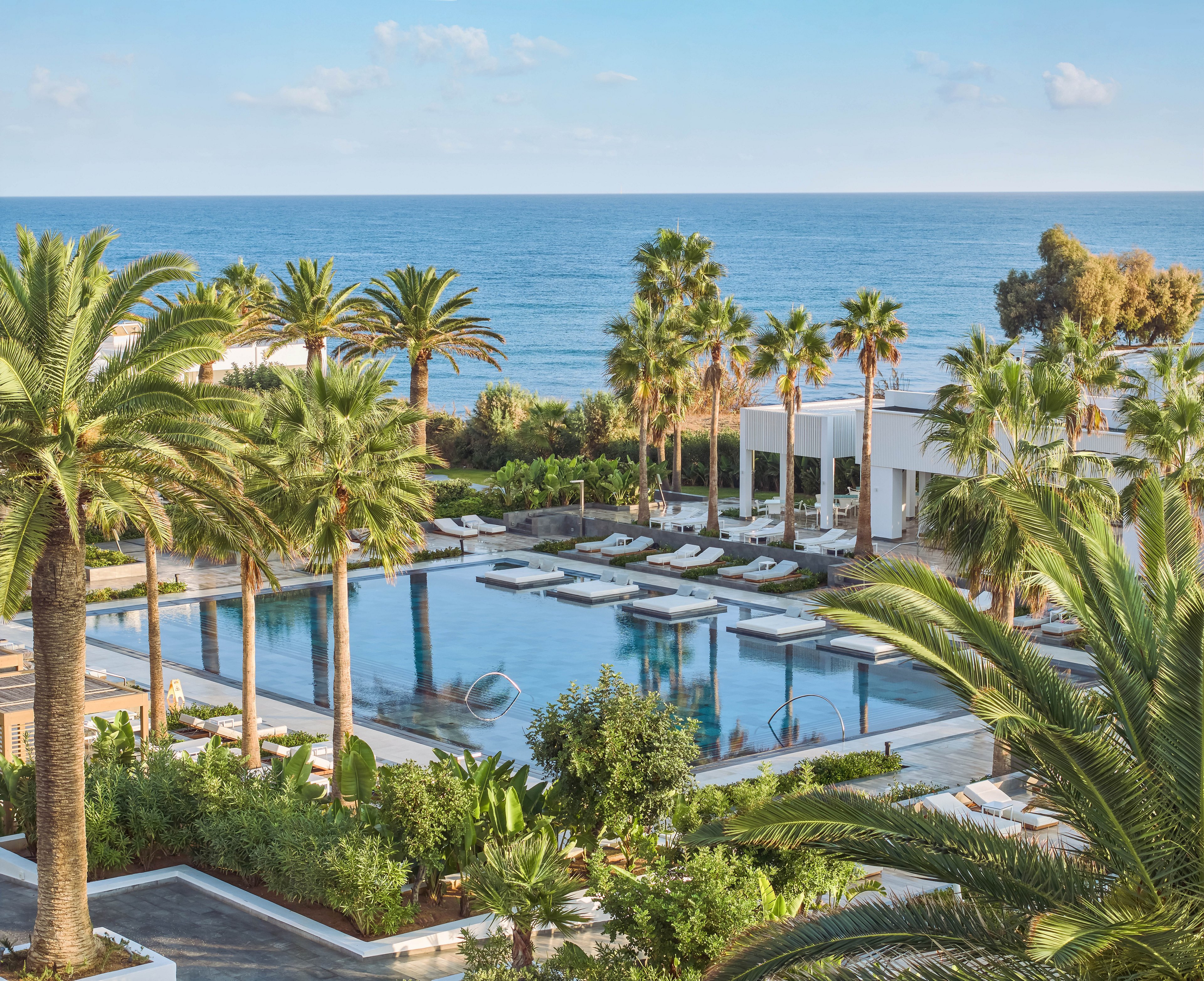 A luxurious resort pool is surrounded by tall palm trees and lush greenery overlooking a calm blue sea. White sun loungers and umbrellas line the poolside, creating an inviting relaxation area. Modern white buildings with open terraces frame the pool, enhancing the clean, minimalist architecture. The clear sky and gentle ocean horizon contribute to a serene, upscale vacation atmosphere.