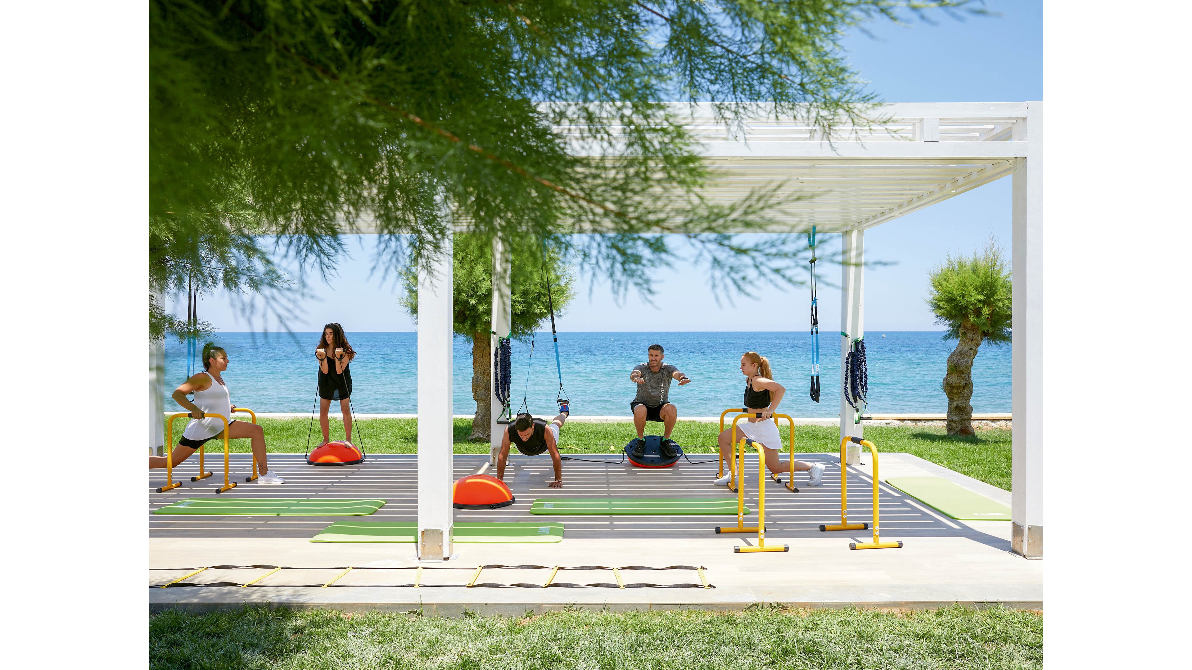 A group of adults participates in an outdoor fitness class under a white pergola near the sea. They use various exercise equipment including balance trainers, resistance bands, and parallel bars. The bright, sunny setting features green grass, trees, and a clear blue ocean in the background. The scene conveys an energetic, healthy lifestyle in a coastal environment.