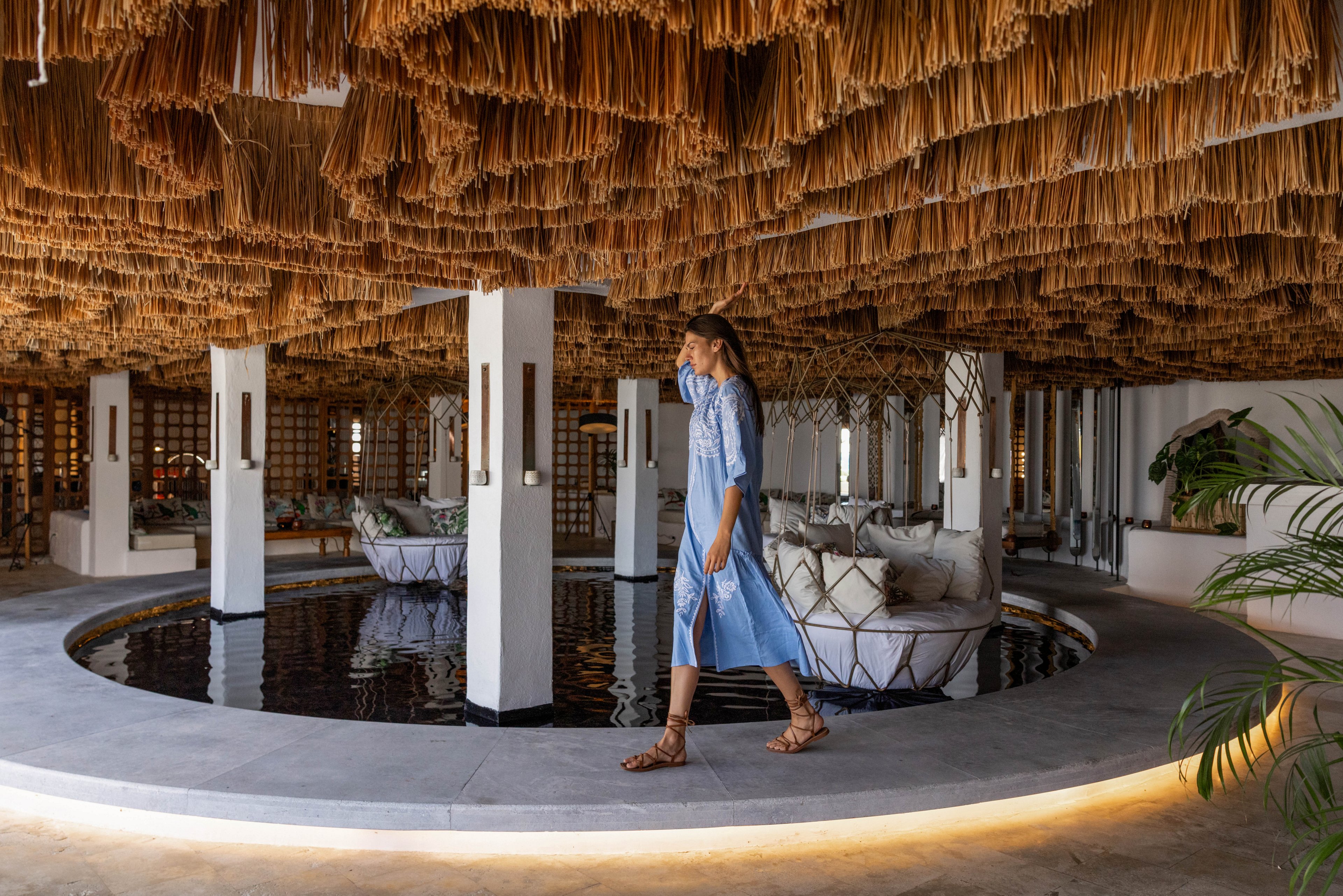 A young woman in a light blue dress walks past a circular indoor water feature in a contemporary lounge. The space features dramatic hanging straw or reed ceiling decor and white structural columns. Cushioned seating areas with neutral-toned pillows surround the pool, creating a relaxed, luxurious atmosphere. Soft, warm lighting accentuates the architectural details and tranquil resort-like setting.