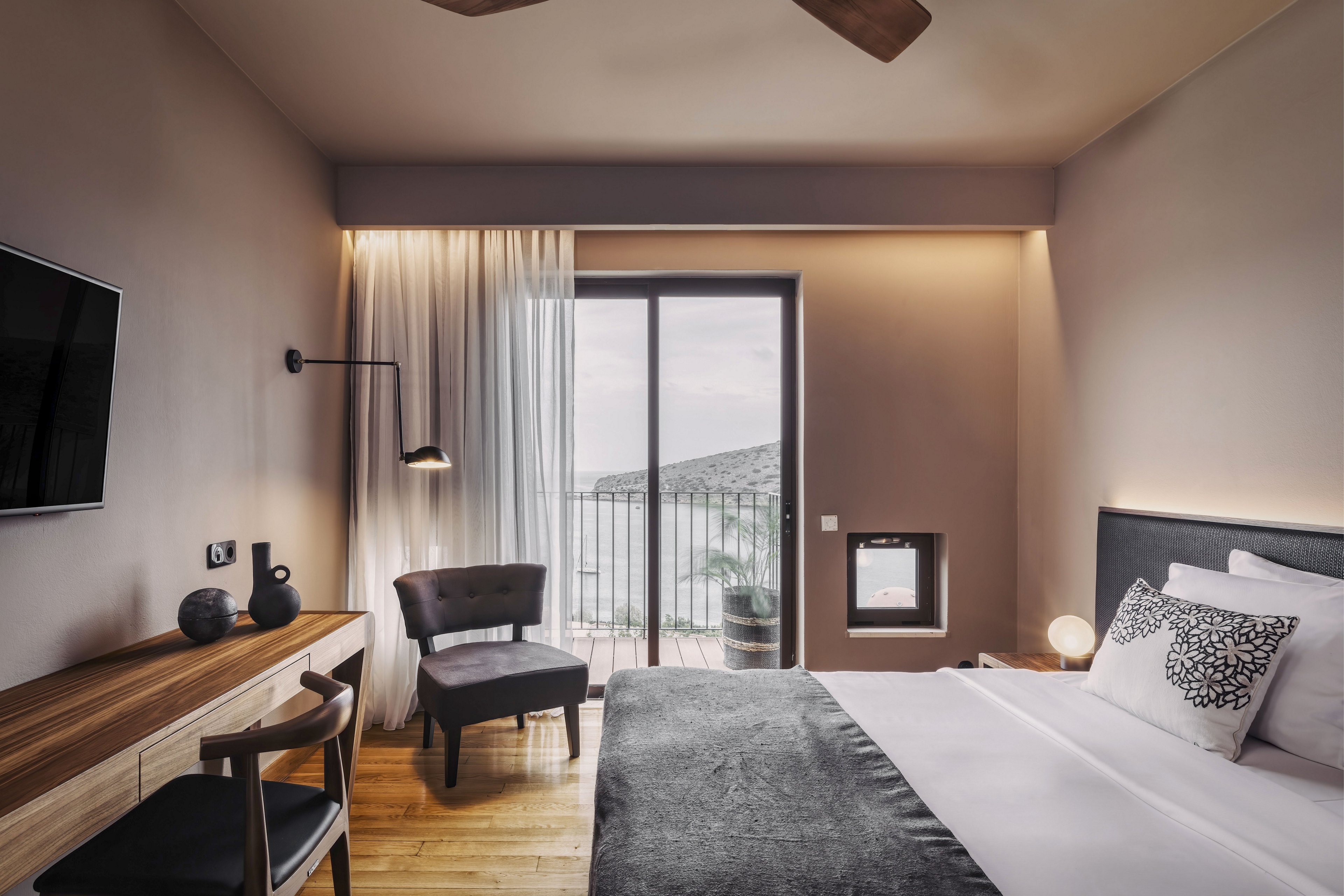 Contemporary hotel room features a neatly made double bed with gray and white bedding. A wooden desk, upholstered chair, and wall-mounted television create a comfortable, functional space. Floor-to-ceiling curtains frame a glass door leading to a balcony with an outdoor view. Warm lighting, neutral walls, and wooden flooring contribute to a calm, minimalist atmosphere.
