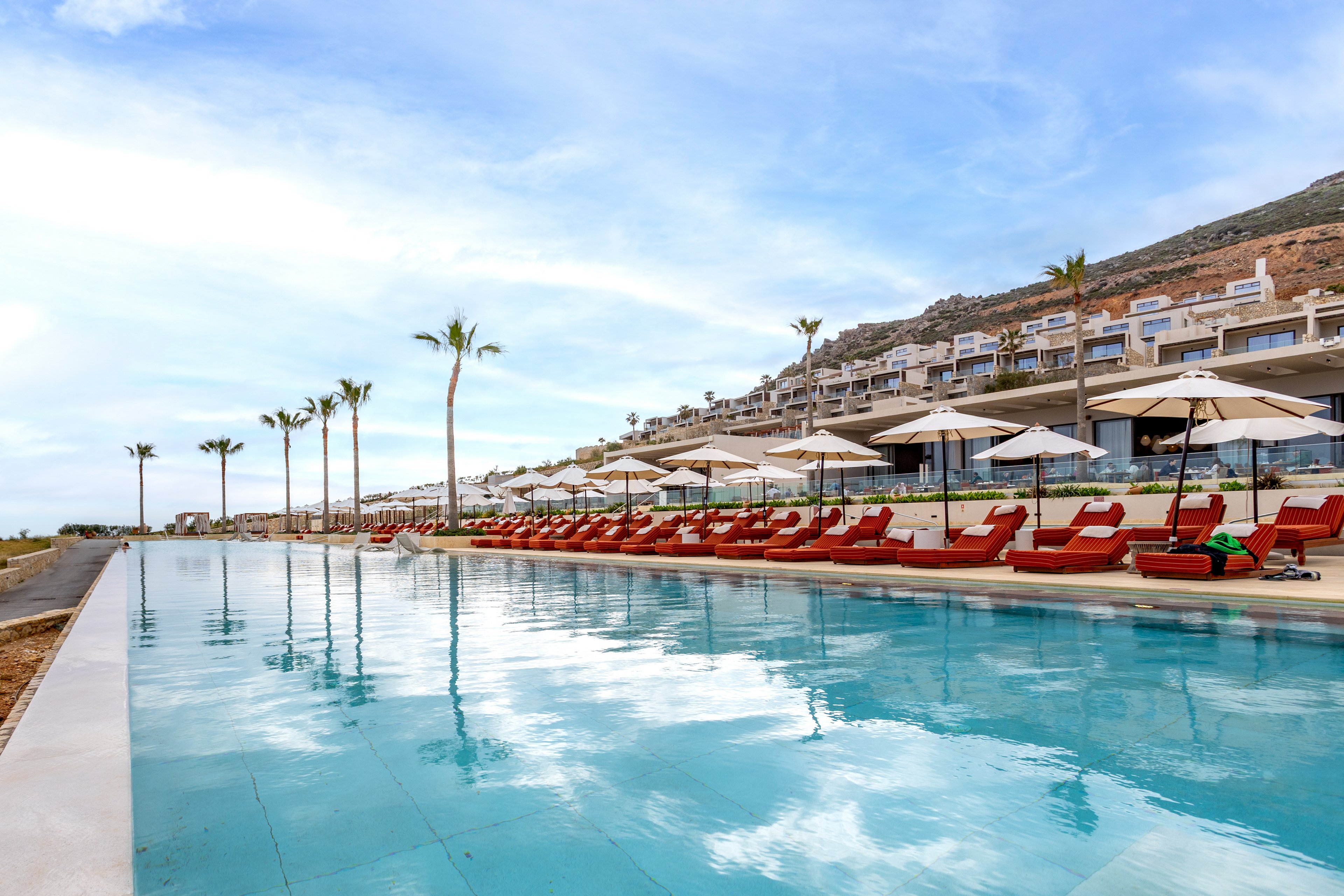 A long infinity pool stretches alongside a modern hillside resort. Rows of red lounge chairs and white umbrellas line the pool deck, facing the water. Palm trees are spaced evenly along the edge, adding a tropical feel to the scene. The sky is partly cloudy, and the calm water reflects the resort buildings and palm trees.
