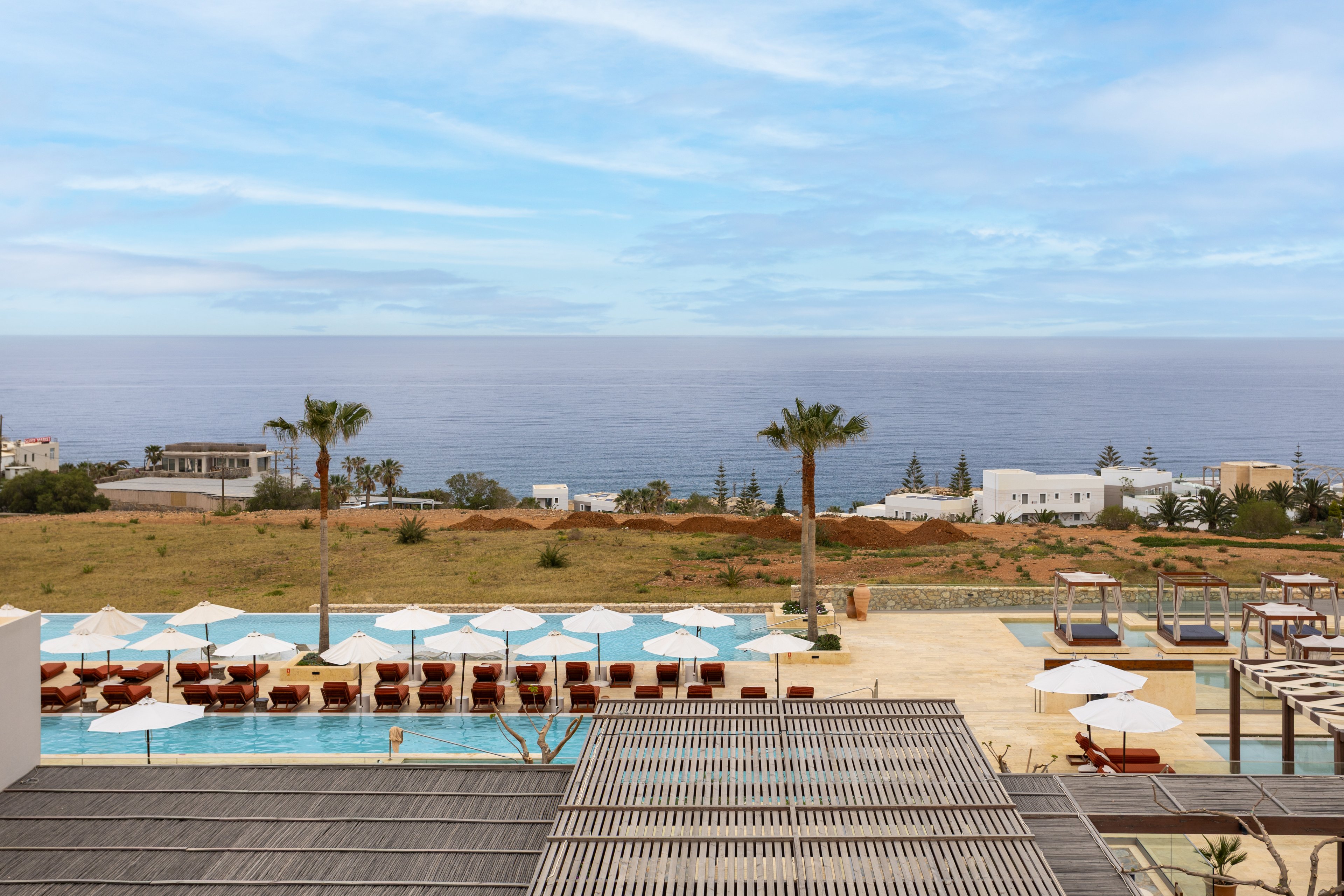 A modern seaside resort features multiple swimming pools lined with rows of white sun umbrellas and lounge chairs. The scene overlooks a grassy coastal landscape leading to calm ocean waters under a partly cloudy sky. Palm trees and low white buildings are visible in the background, adding to the Mediterranean-style setting. The overall atmosphere is tranquil and inviting, ideal for a relaxing vacation by the sea.