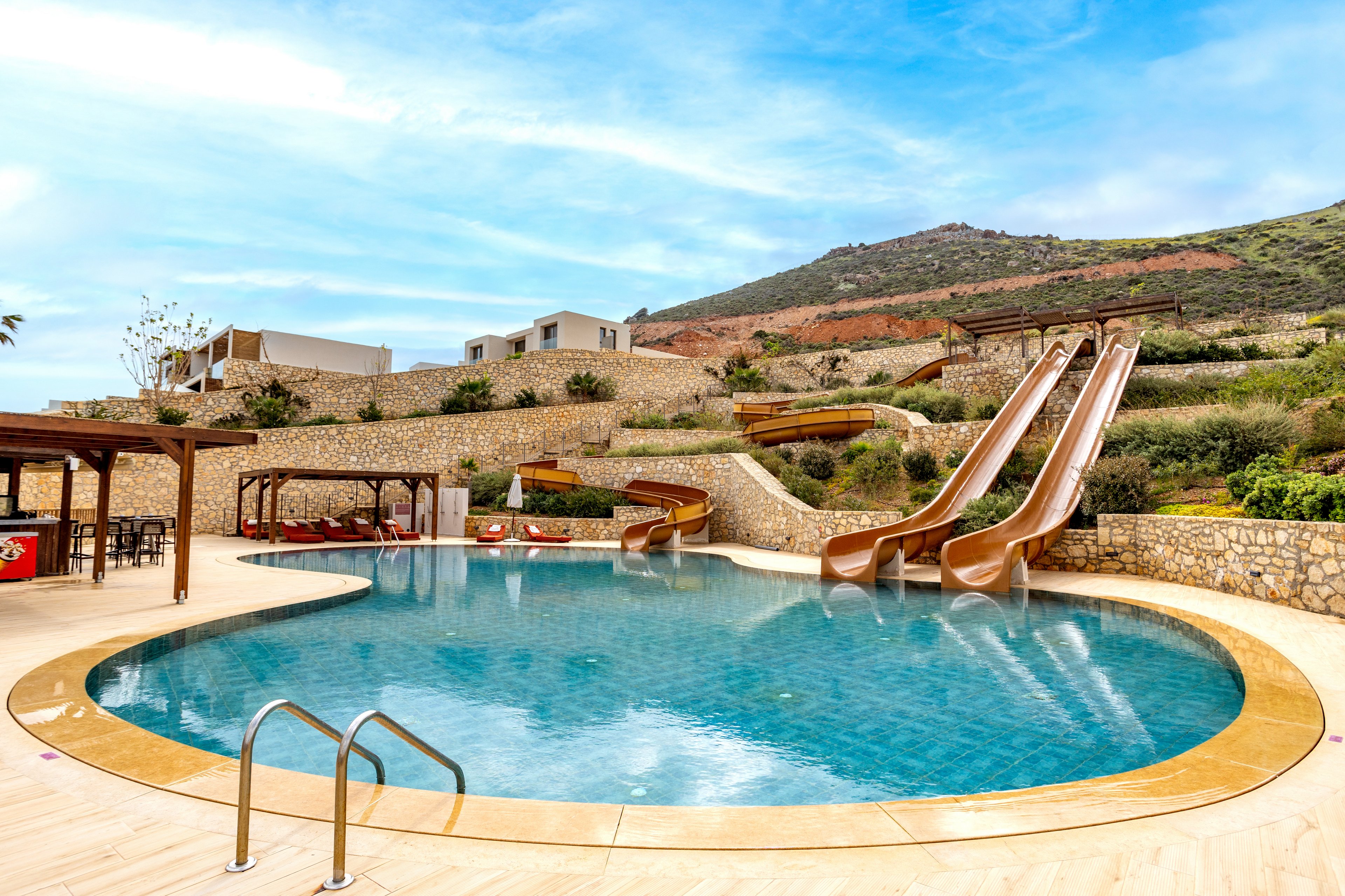A clear blue resort swimming pool sits in the foreground with metal steps leading into the water. Several tall waterslides curve down a landscaped hillside into the pool area. Stone terraces, greenery, and modern white buildings create a layered backdrop against a bright blue sky. Wooden pergolas with shaded seating and lounge chairs add to the relaxed vacation atmosphere. The scene is calm and inviting, with no people present.